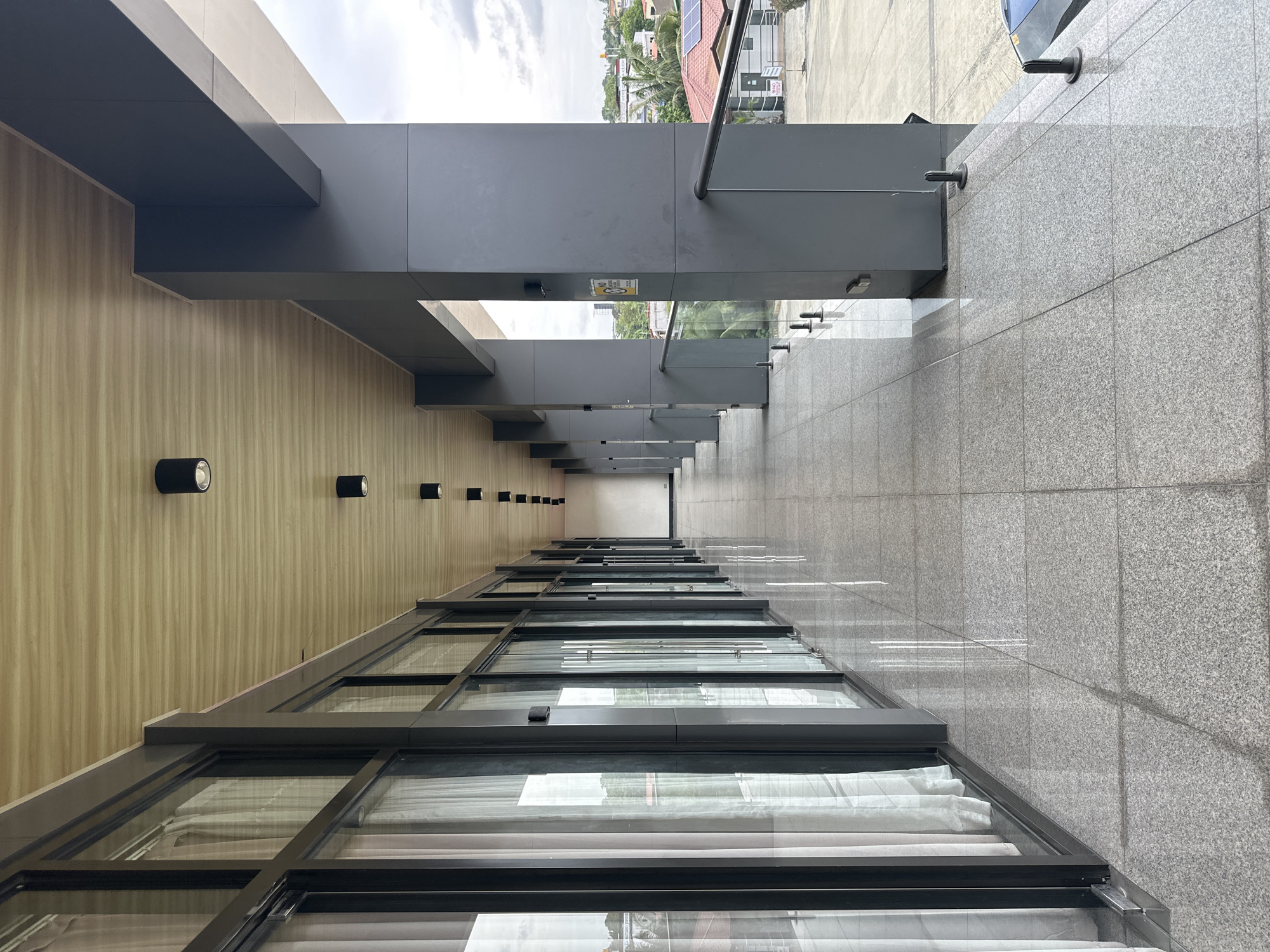 The image captures a long, modern, covered outdoor walkway or hallway that appears to be located in a commercial or residential building in Davao City, Philippines. The walkway features a polished granite tile floor that reflects the overhead lights and the adjacent building's exterior. On the left side, a series of floor-to-ceiling glass windows with white curtains are visible, suggesting interior rooms or offices. The ceiling is constructed of wooden planks with evenly spaced black cylindrical lights. On the right side of the walkway, there are large, dark gray square columns supporting the overhanging structure, with a glass railing system demarcating the edge of the walkway and offering a view of the surrounding urban landscape. This landscape includes various buildings, some with red tile roofs, trees, and a cloudy sky. A yellow warning sign is attached to one of the columns. The overall impression is one of a well-maintained, contemporary architectural space within a tropical city setting.