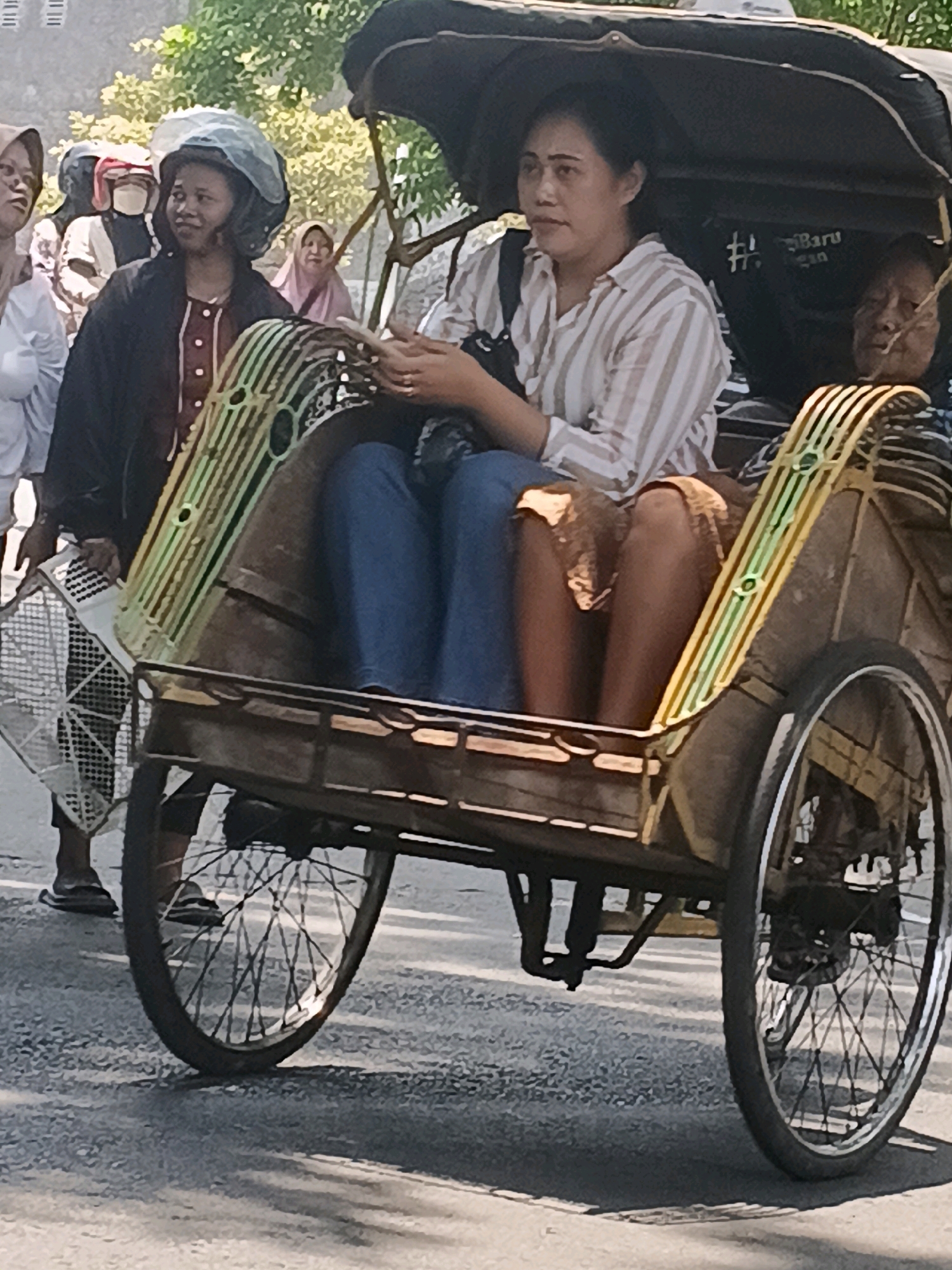The image shows a pedicab transporting passengers in Purwodadi, Indonesia. In the pedicab, two women are seated. The woman in front, wearing a striped shirt and skirt, is looking forward. Behind her, another woman is partially visible, wearing a dark headscarf. A third woman, wearing a black jacket and a helmet, stands near the front of the pedicab, looking to the left. Two other women are visible in the background on the left, and the scene appears to be on a street with trees and buildings.
