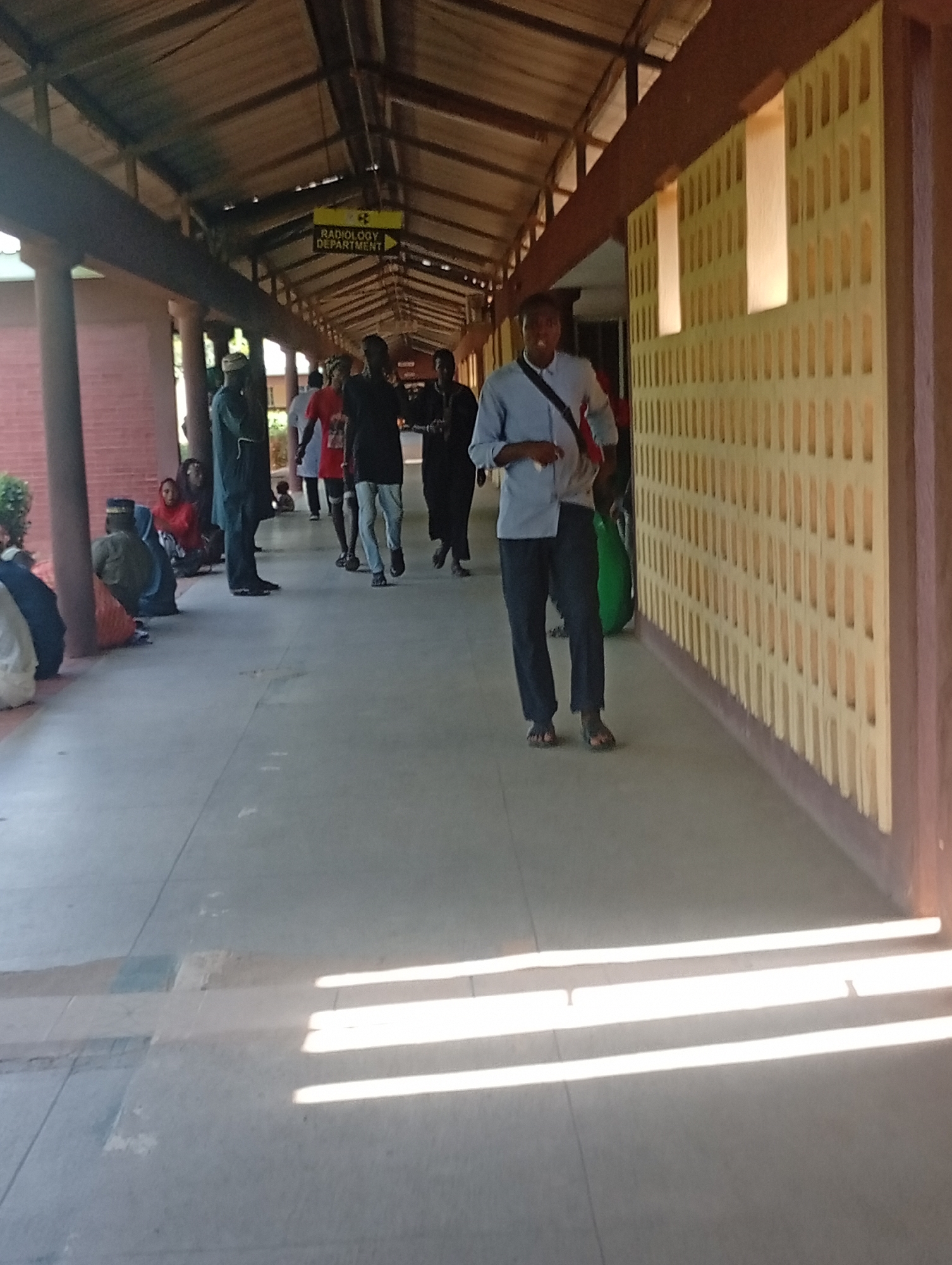 The image depicts a covered walkway in Gombe, Nigeria. A sign indicating "Radiology Department" is visible overhead. Several people are present, some walking and others seated along the side of the walkway. The architecture suggests a public building, possibly a hospital or clinic, with brick walls and a wooden-beamed roof. Sunlight streams through openings in the wall on the right side of the image, casting distinct shadows on the ground.