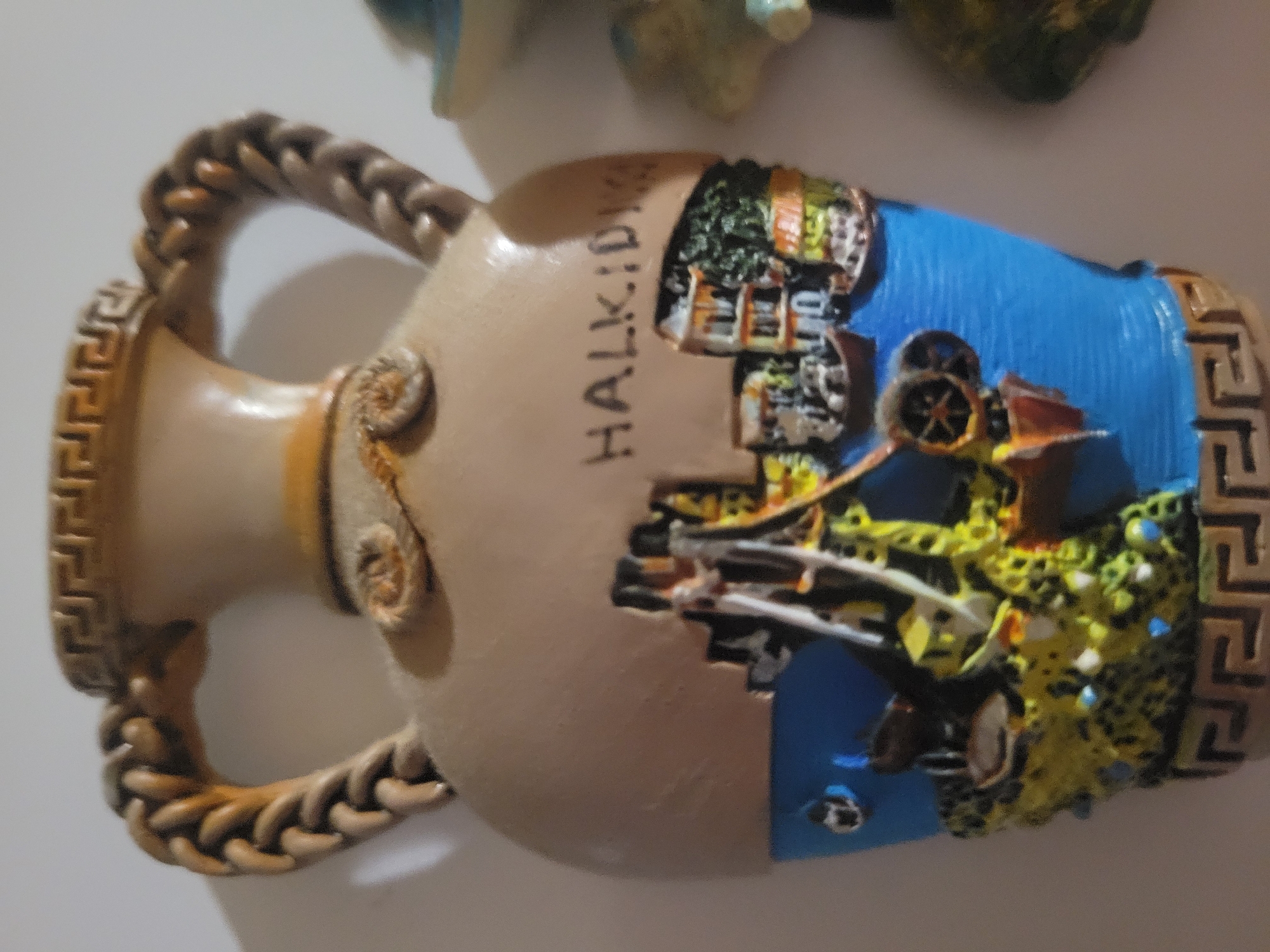 The image shows a close-up of a decorative ceramic amphora. The amphora is beige with a Greek key pattern around the rim and base. It has two braided handles. The body of the amphora is decorated with a relief scene depicting a waterfront with buildings, boats, and lush greenery. The word "HALKIDIKI" is written in black lettering on the upper part of the amphora. The scene is detailed and colorful, with vibrant blue for the water and bright green and yellow for the landscape. The overall impression is that of a souvenir or decorative item. No location in Belaazyorsk, Belarus is depicted.