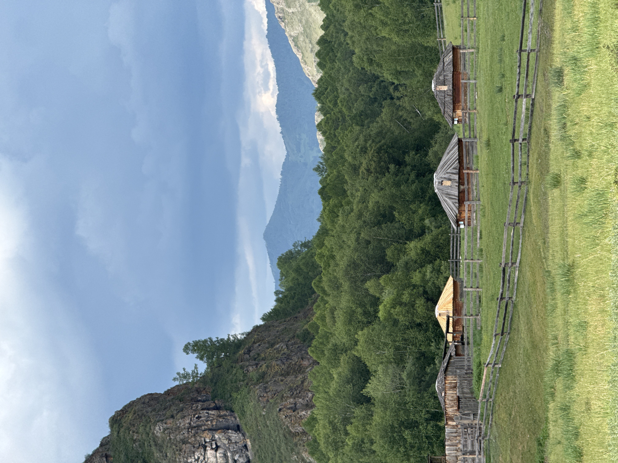 The image depicts a rural scene in the village of Kazanovka, Russia. In the foreground, a wooden fence encloses a grassy field. Behind the fence, several traditional wooden structures are visible, resembling yurts or roundhouses with thatched roofs. One of these structures has a bright yellow, conical roof. A larger wooden building with a sloped roof is situated to the left of these. The middle ground is dominated by a dense forest of green trees. In the background, majestic mountains rise, with the closest ones covered in trees and the further ones displaying rugged, rocky slopes. The sky above is a mix of gray and white clouds. The scene suggests a tranquil, natural landscape with elements of traditional architecture.