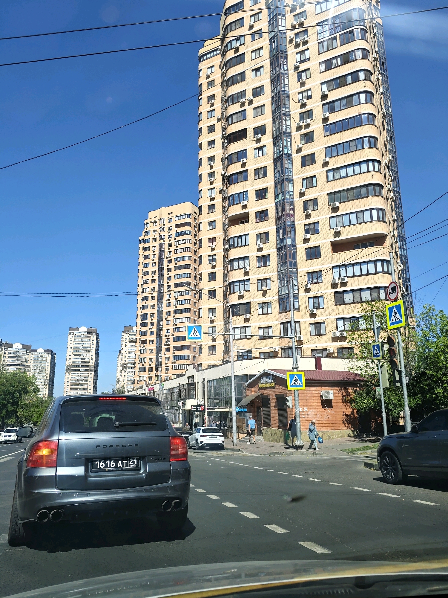 The image depicts a street scene in Rostov-on-Don, Russia, on a clear, sunny day. In the foreground, the rear of a dark gray Porsche Cayenne SUV is prominently displayed, its license plate reading "1616 AT 21 RUS". The SUV is in motion or stopped at traffic. Across the street, several multi-story buildings with light beige facades and numerous windows dominate the background, characteristic of modern urban architecture. Some of these buildings appear to be residential complexes, with visible air conditioning units attached to their exteriors.
To the right of the Porsche, a traffic light and pedestrian crossing signs are visible, indicating a regulated intersection. Several people are seen walking on the sidewalk near a small brick building with a sign that appears to say "БОЧКА" (BOCHKA), suggesting a local shop or establishment. Green trees line the side of the road, adding a touch of nature to the urban landscape. The sky is a vibrant, cloudless blue. The overall impression is of a typical city street on a pleasant day, with everyday urban activity in progress.