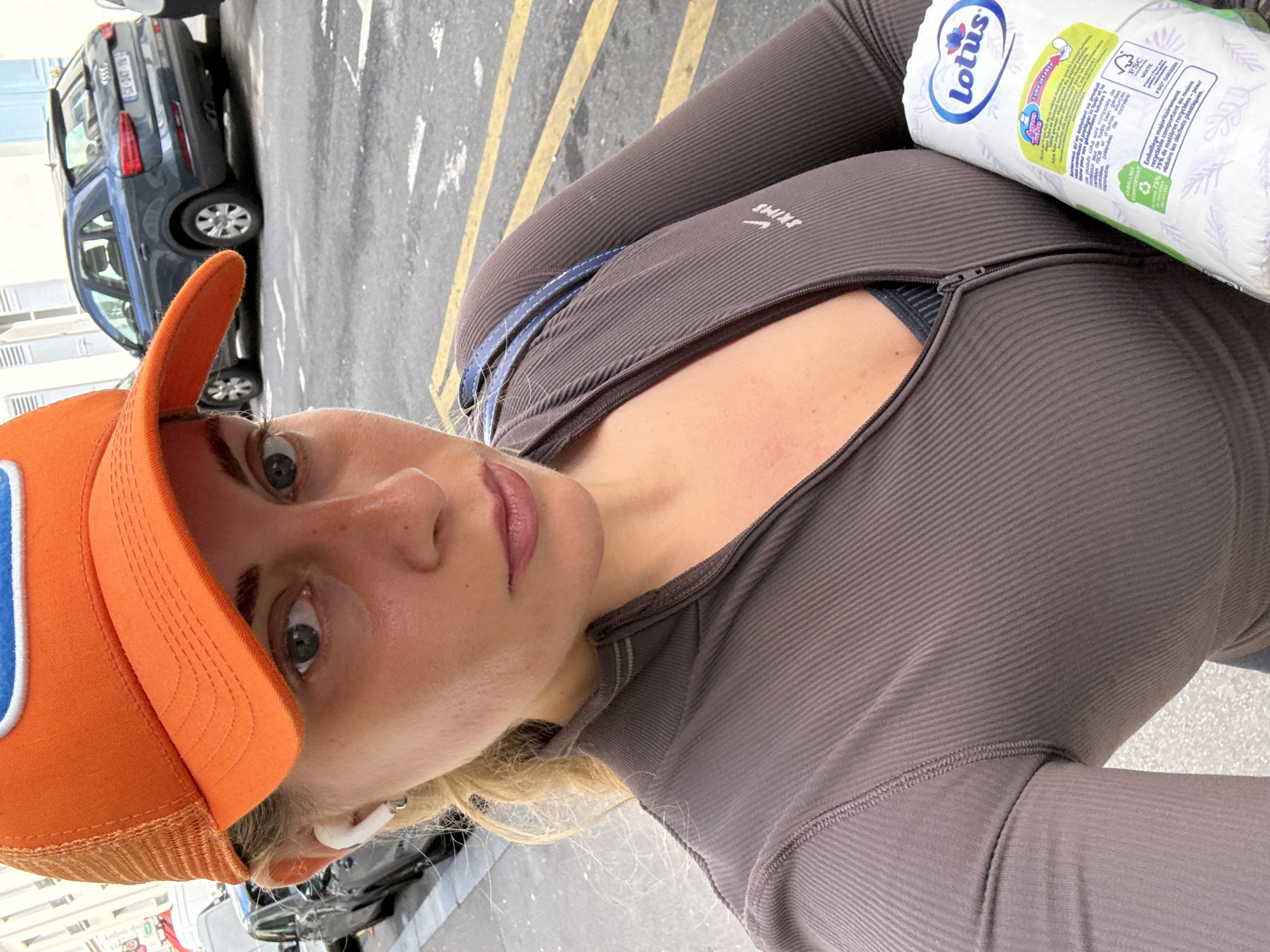 A selfie of a woman in Paris, France. She is wearing an orange baseball cap, a brown ribbed zip-up top with an unzipped front, and earbuds. She is looking at the camera with a neutral expression. She is holding a package of Lotus brand toilet paper against her chest. Behind her, a vehicle is parked near a street with yellow striped markings. The buildings are white, and the weather is sunny.