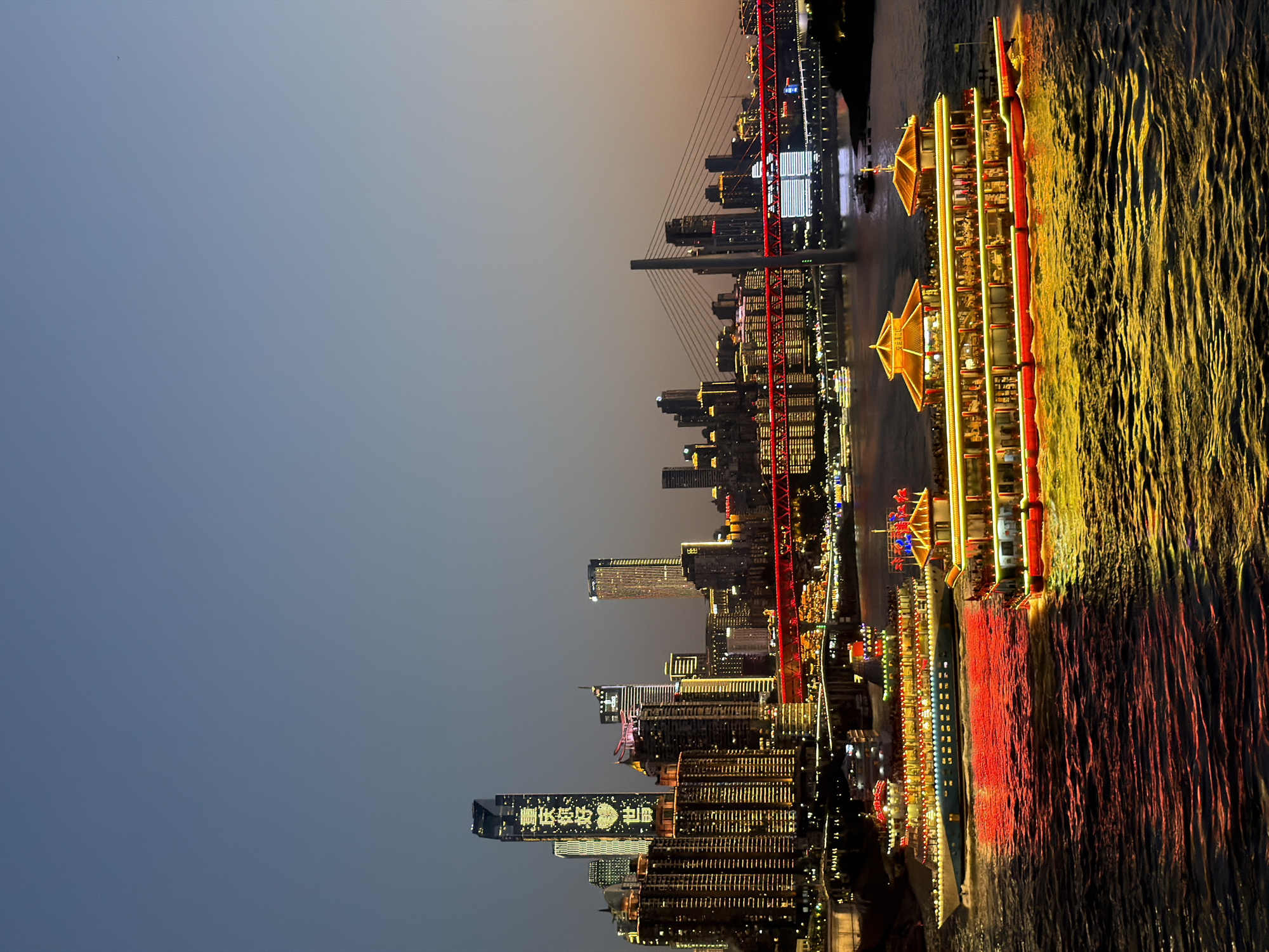 This is a vertical, eye-level, full shot of the Nan'an District in Chongqing, China at dusk. The scene is dominated by the cityscape with numerous illuminated buildings reflecting on the water's surface. Two tour boats are prominently featured in the foreground, emitting bright red and yellow lights that cast colorful reflections on the rippling water. The boat on the right is larger and has a more ornate, pagoda-like structure on its roof, adorned with traditional Chinese architectural elements and golden accents. The boat on the left is longer and more modern in design, also brightly lit. In the background, a suspension bridge with red lighting spans the river, connecting the Nan'an District to another part of the city. Beyond the bridge, a cluster of tall skyscrapers forms the city skyline, with many buildings illuminated by golden lights. One particularly tall skyscraper in the center of the skyline displays a message in bright yellow lights, appearing to be Chinese characters. The sky above is a gradient of dusky blue and orange, indicating the transition from day to night. The water appears choppy, with distinct wave patterns and reflections. There are no visible people in the image, as the focus is on the illuminated cityscape and the boats.
