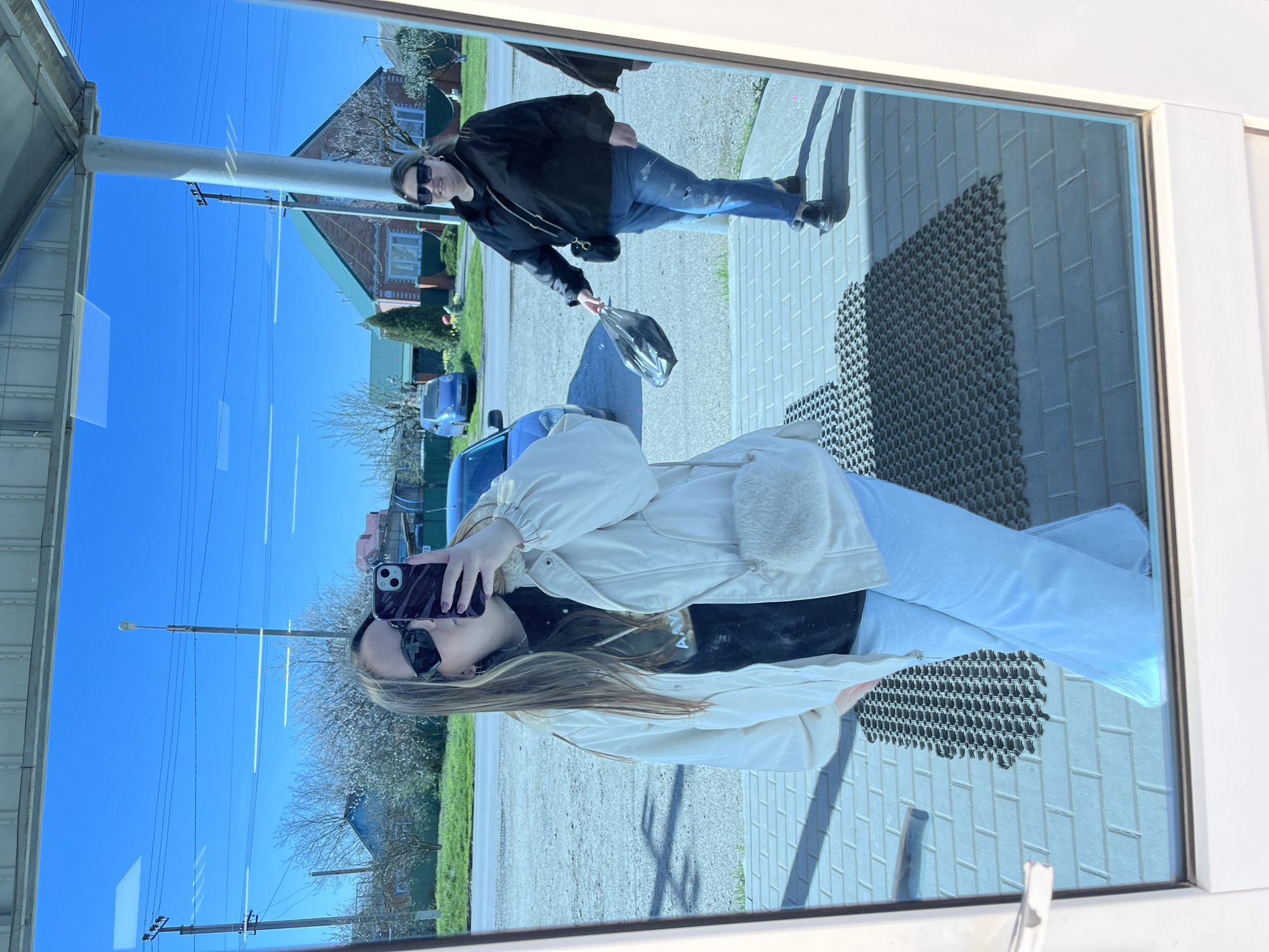 A photo shows a reflection in a glass window. A woman in a light-colored jacket and jeans is taking a picture of herself with a cell phone. Reflected in the background is a street scene in Novopokrovskaya, Russia, featuring a blue car, a two-story house with a green roof, utility poles, and another person walking on the sidewalk wearing a black jacket and jeans. The foreground includes a door frame and a dark patterned floor mat.