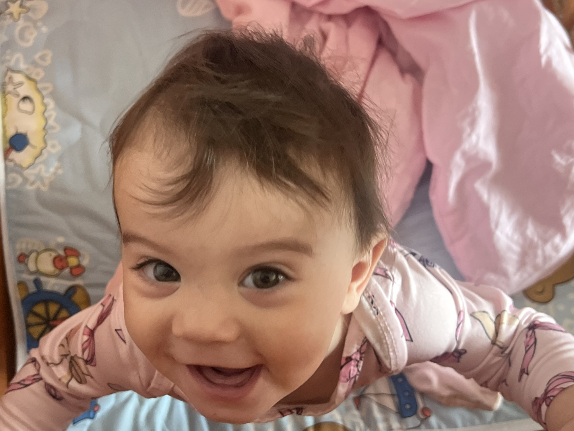 A baby lies on a blue patterned sheet with a pink blanket behind them. The baby has brown hair and eyes, and is smiling with their mouth open. The baby is wearing a pink patterned long-sleeved shirt. The sheet has drawings of stars, moons, a clock, and a sun on it. The location in the city of Elena, Bulgaria is not discernable from the image.