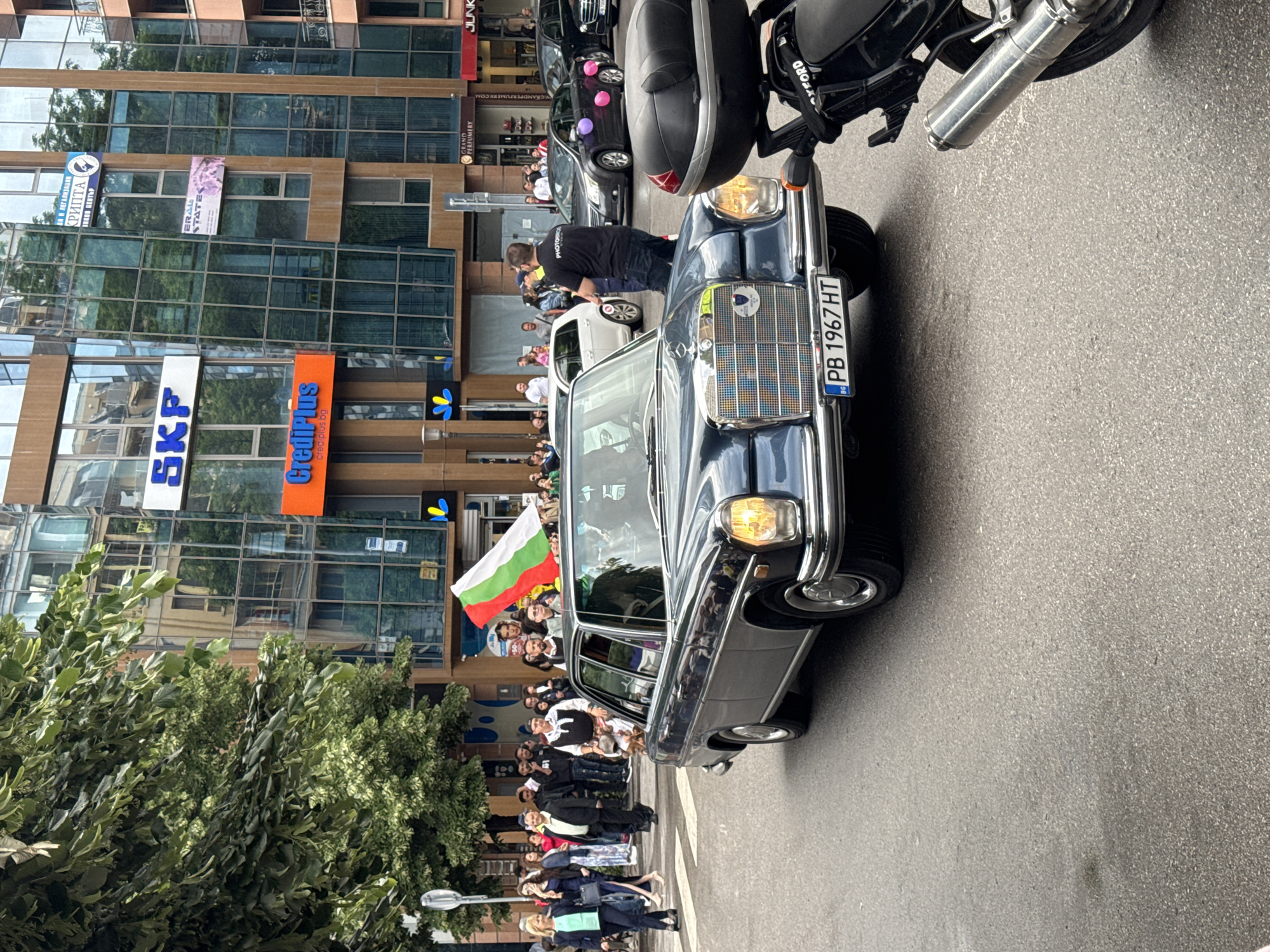 The image shows a street scene in Plovdiv, Bulgaria. An older, dark-colored Mercedes Benz car is in the foreground, with a Bulgarian flag waving from it. A crowd of people stands along the sidewalk, some of whom appear to be waving or observing the car. In the background, there is a modern building with glass windows, displaying signs for "SKF" and "CrediPlus." Another sign reads "Photobro," and another is "ERALL STATES" (unreadable). A license plate on the car reads "PB 1967 HT." Part of a motorcycle is visible in the upper right corner of the frame. A tree and a street lamp are also visible on the left side of the scene. Other cars and people can be seen behind the Mercedes.