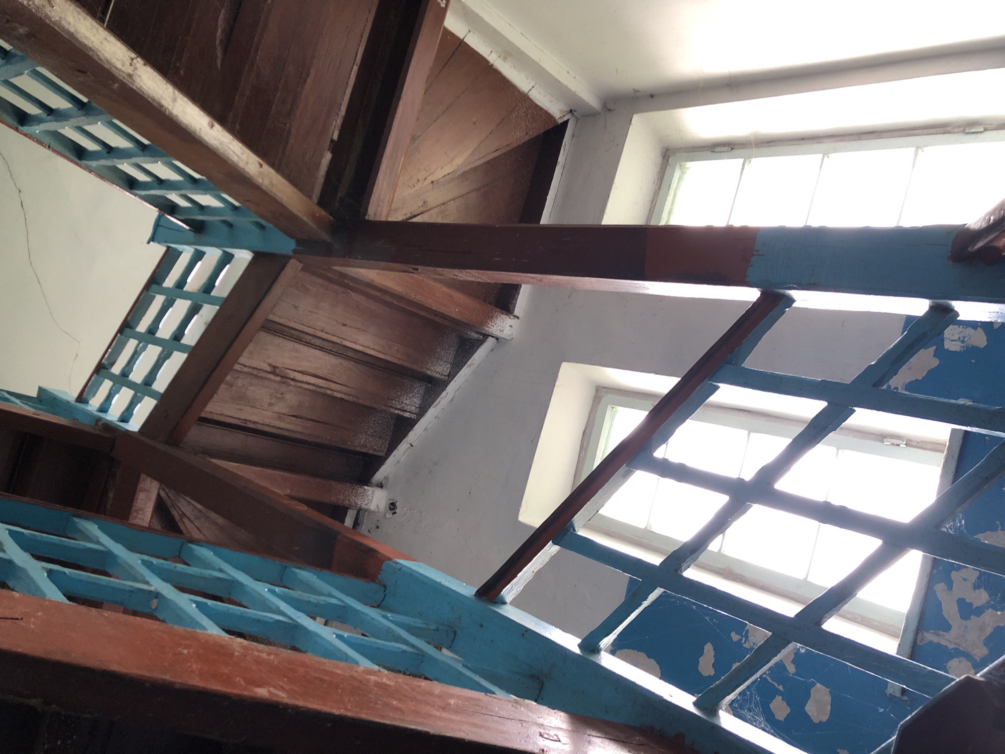 The image shows a view looking up at a wooden staircase with blue and red painted railings. The staircase has multiple levels visible, with wooden beams and planks forming the ceiling and supports. There are windows on either side, with bright light coming through, obscuring details outside. Cobwebs are visible on the blue painted surfaces and in some of the wooden structures. The walls are painted white. The overall impression is of an older, possibly dilapidated interior space.