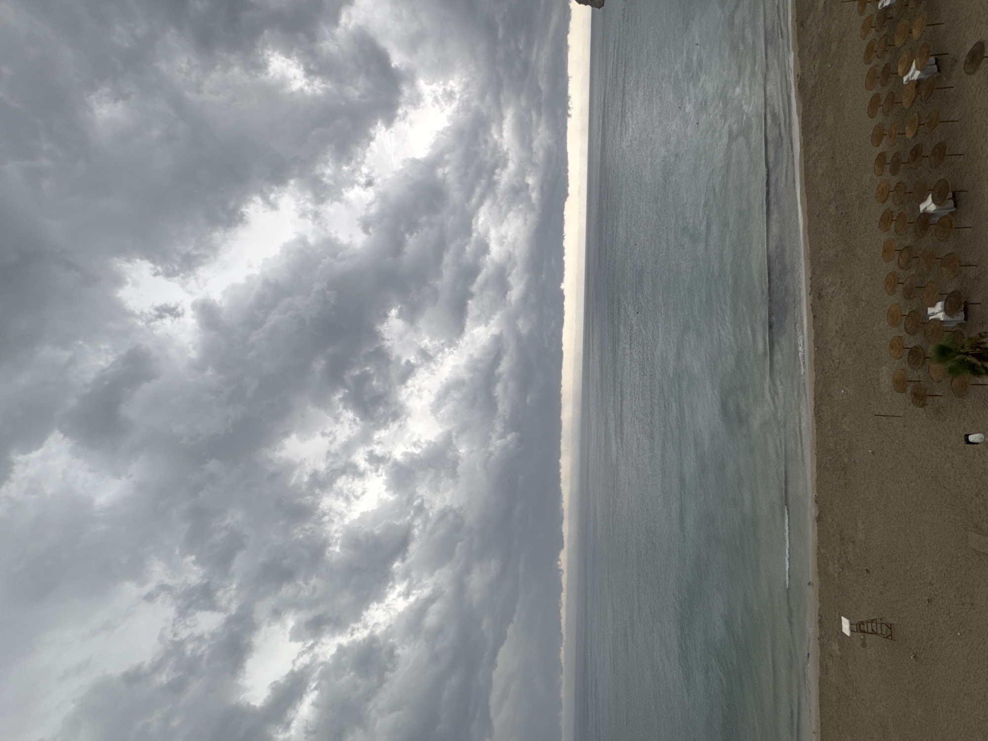 This image depicts a beach in Calp, Spain, under a heavy, overcast sky filled with dark, dramatic clouds. The sea is a muted gray-blue, with small waves lapping at the sandy shore. On the beach, there are rows of closed straw umbrellas, some with chairs underneath. A red lifeguard stand with a white canopy is situated on the sand near the water's edge. A small palm tree is visible on the far left of the beach. The scene conveys a somber and potentially stormy atmosphere, typical of a cloudy day at the coast.