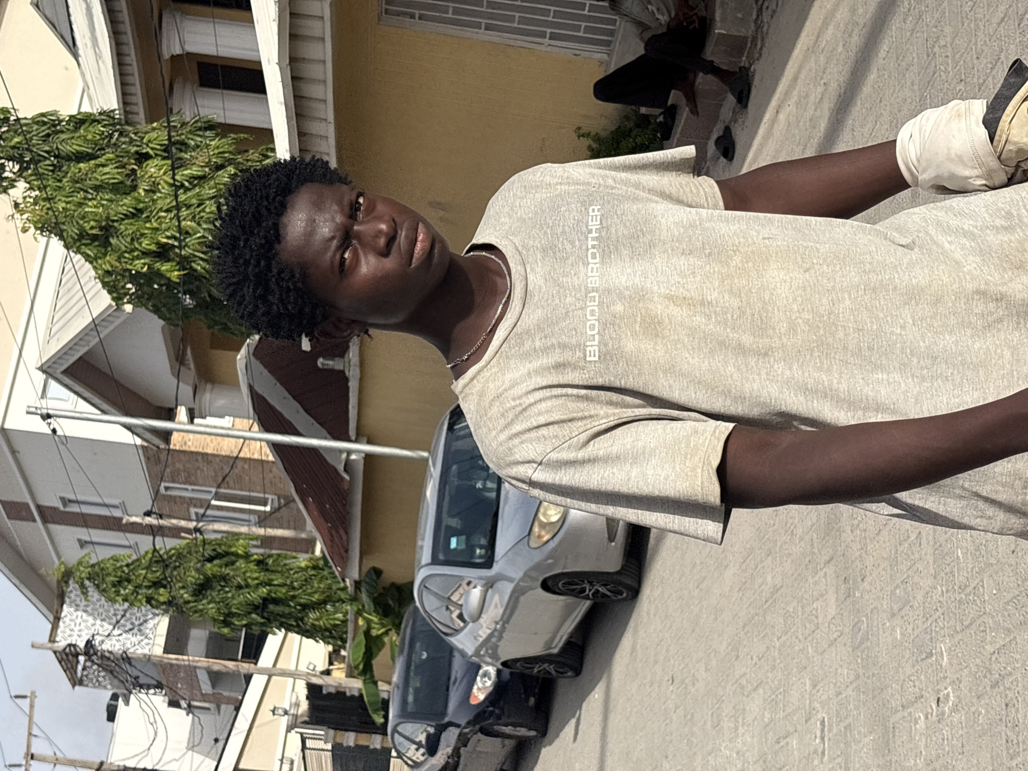 A medium, angled outdoor shot shows a dark-skinned young man in Igboefon, Nigeria, wearing a gray t-shirt with "BLOOD BROTHER" printed vertically on the chest, a silver chain, and a dirty white athletic glove on his right hand. He has short, dark curly hair. He is standing on a street. Behind him are parked cars and residential buildings with trees.