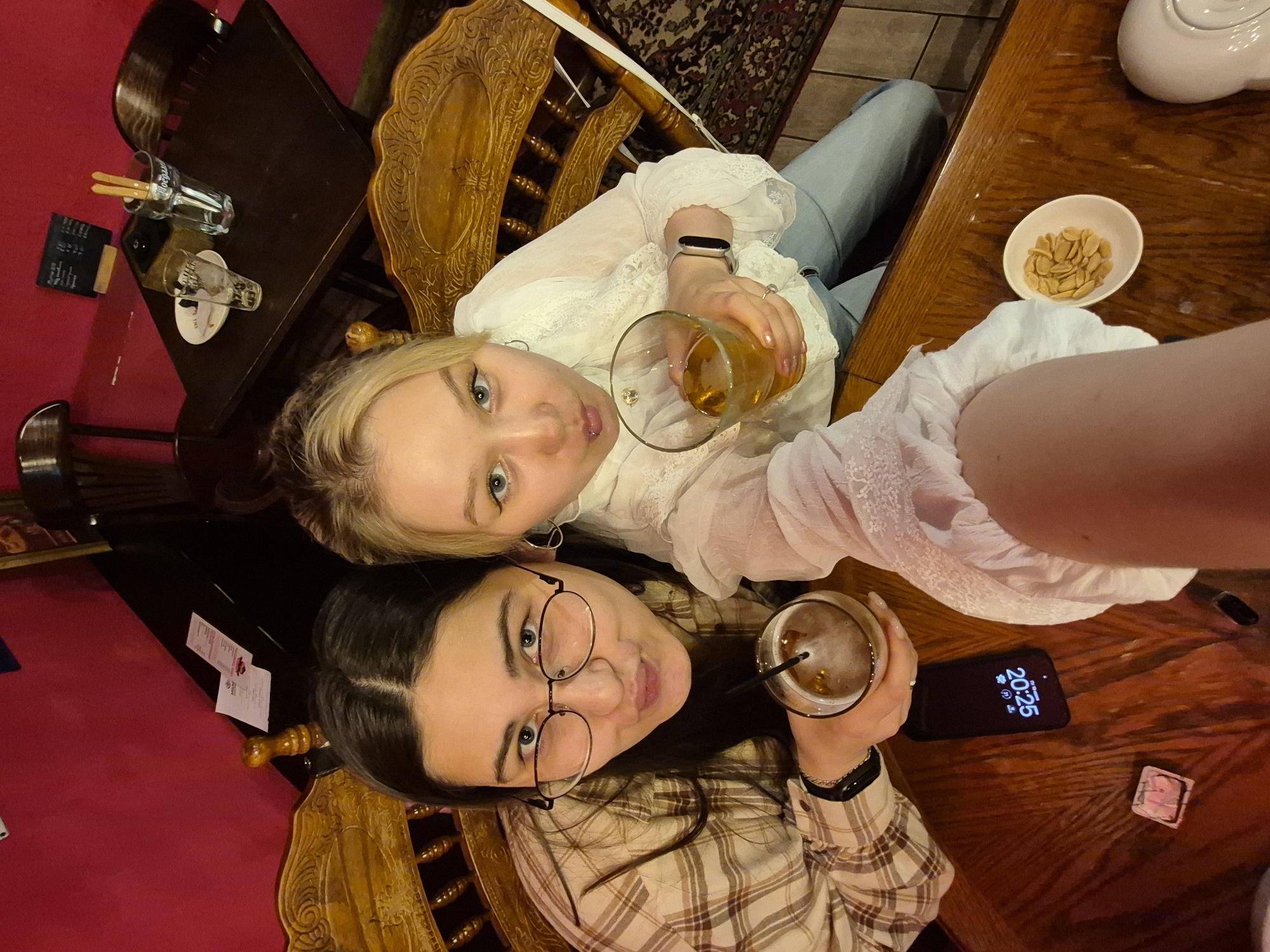 The image captures a high-angle, selfie-style shot of two young women seated at a wooden table in what appears to be a cafe or bar in Chaikovsky, Russia. The woman on the left, wearing a plaid shirt and glasses, holds a drink with a straw. The woman on the right, in a white lace blouse, holds a glass of amber liquid. A smartphone displaying the time 20:25 is on the table between them, along with a small dish of nuts and what appears to be a receipt. The background features ornate wooden chairs and a vibrant red wall. The overall ambiance suggests a casual social gathering.