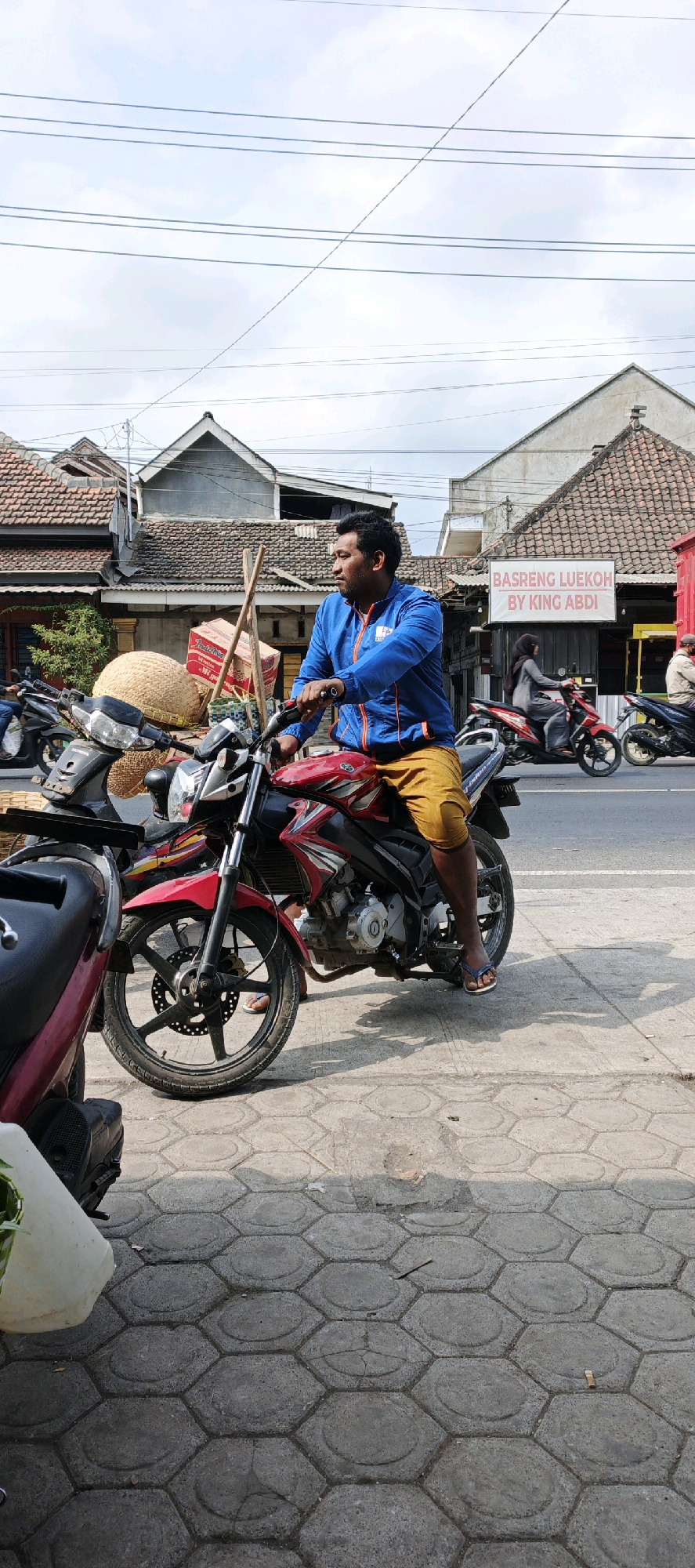 A man wearing a blue jacket and yellow shorts sits on a red motorcycle on a paved sidewalk in Kasiyan, Indonesia. The motorcycle is loaded with goods, including a large woven basket and several boxes, one of which is labeled "Indomie". Behind the man, a sign reads "BASRENG LUEKOH BY KING ABDI". Several other motorcycles are visible on the street and sidewalk, and buildings with tiled roofs line the street. The sky is overcast.