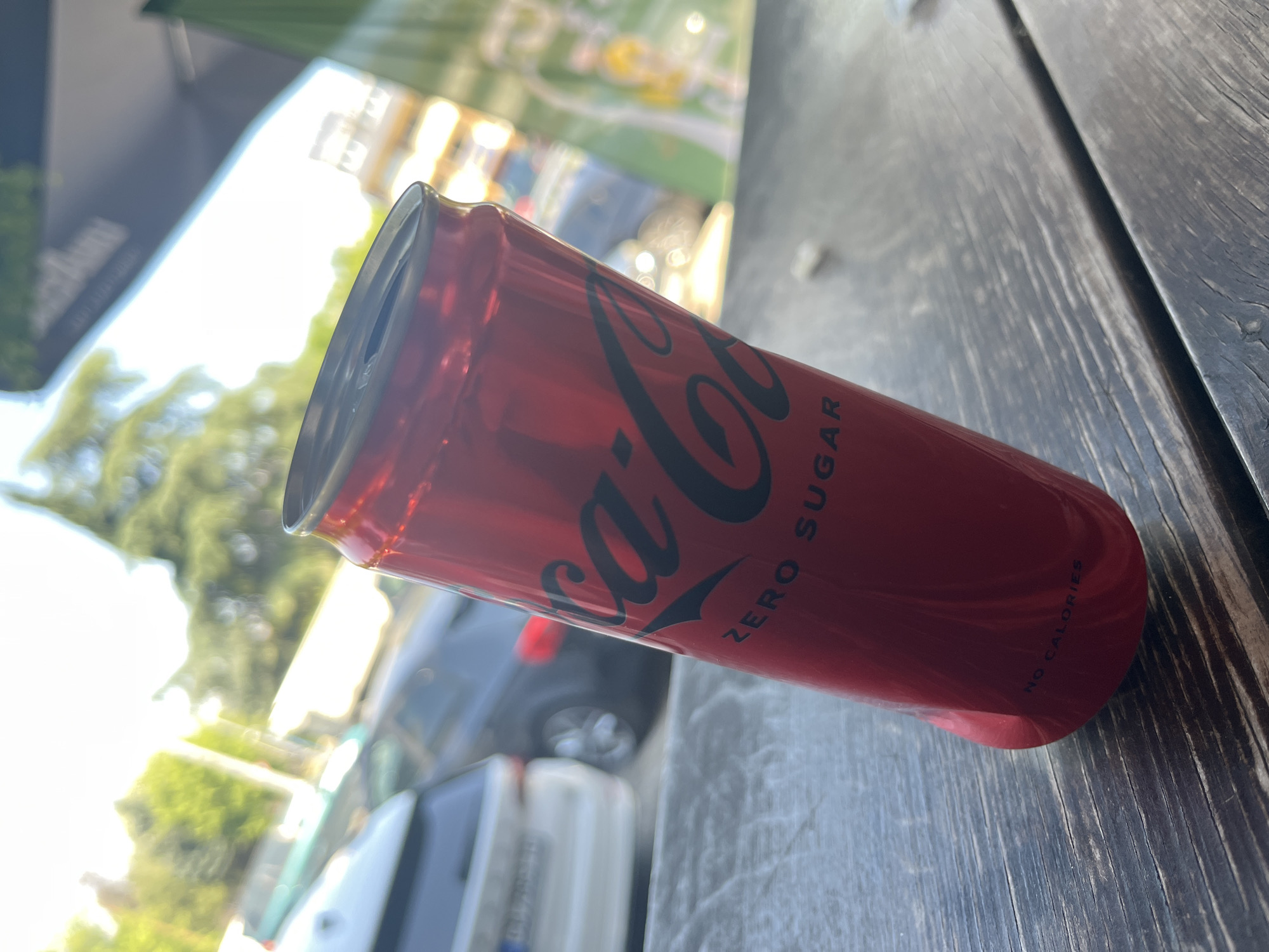 A Coca-Cola Zero Sugar can rests on a weathered, wooden surface, likely a table or bench, in an outdoor setting. The can is primarily red with the iconic Coca-Cola logo in black, along with the text "ZERO SUGAR" and "NO CALORIES." The can is positioned at an angle, revealing its metallic top. The background shows an urban landscape, including vehicles and foliage, suggesting a city environment. The location details, specifically the city of Unknown, Unknown, are unclear due to limited visual information.