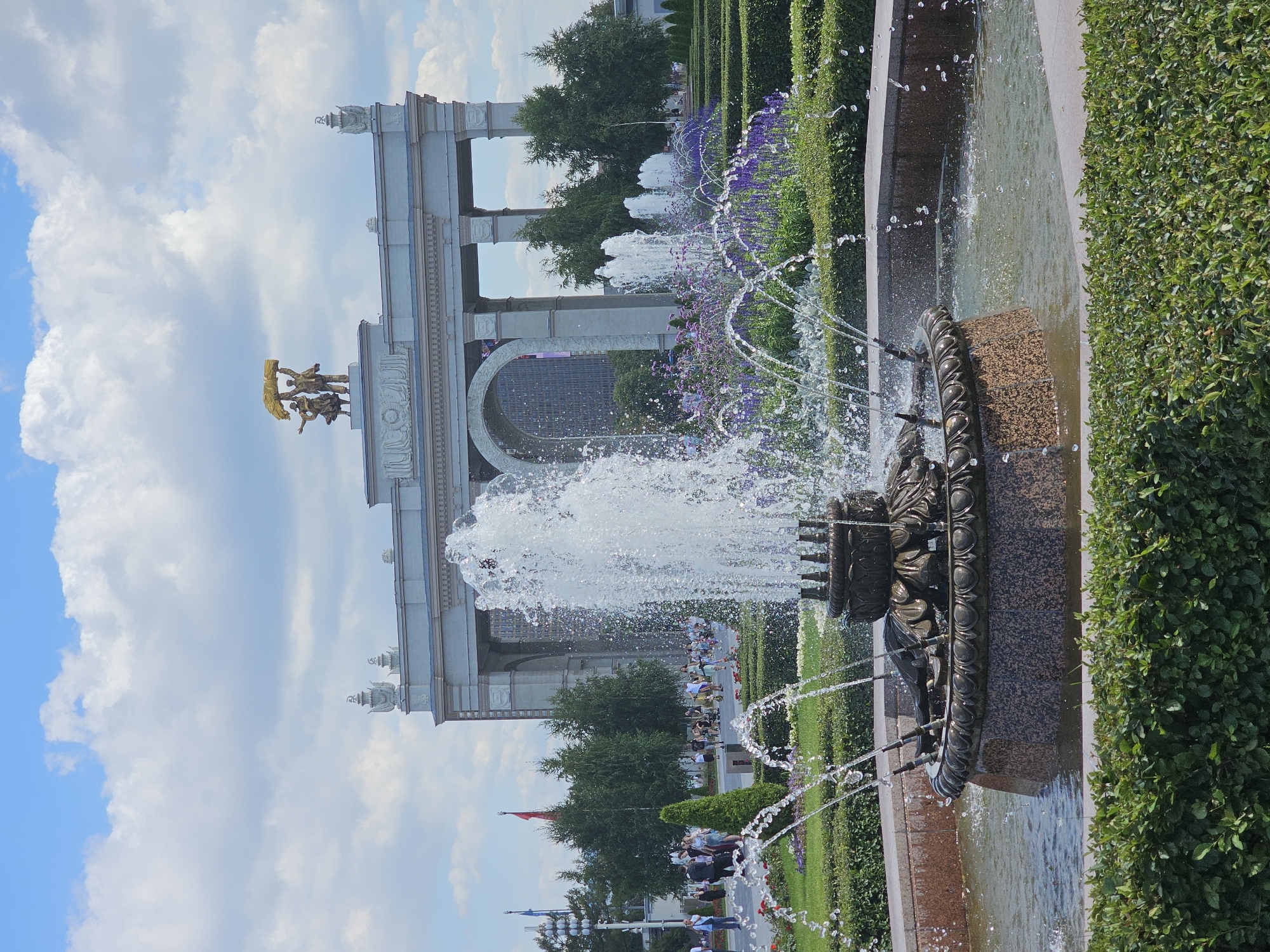 The image depicts a vibrant outdoor scene at the All-Russian Exhibition Centre (VDNKh) in Moscow, Russia, on a partly cloudy day. In the foreground, a multi-tiered bronze fountain with water jets in motion dominates the center of the frame, surrounded by manicured green hedges and flower beds with purple blooms. Behind the fountain, a grand, neoclassical archway with Corinthian columns serves as the backdrop. Atop the archway, a golden statue of two figures holding a sheaf of wheat stands prominently against the sky. The area around the archway features more lush greenery and scattered trees. A crowd of people, dressed in casual summer attire, is visible walking and congregating in the middle ground, some near the fountain and others further back towards the archway and buildings. The overall atmosphere is one of a lively public space on a pleasant day.