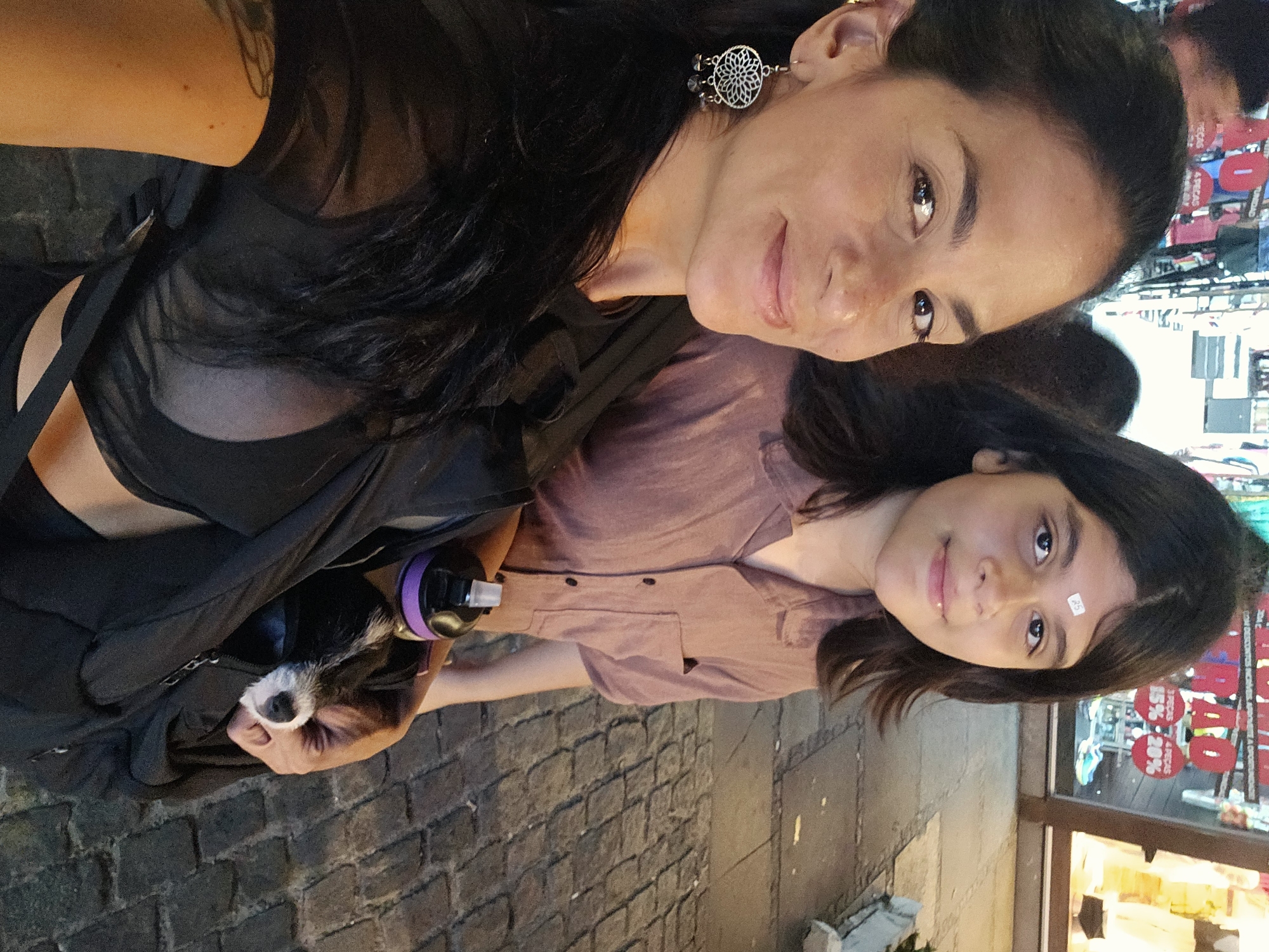 Two women are in a self-portrait photo on a cobblestone street in Armação Dos Búzios, Brazil. The woman in the foreground wears a black shirt with mesh elements and large circular earrings. She carries a black bag with a small dog inside. Next to her, the second woman wears a mauve button-down shirt. There is a small sticker on her forehead. A water bottle with a purple lid is visible next to the dog. A shop window with sale signs is in the background.