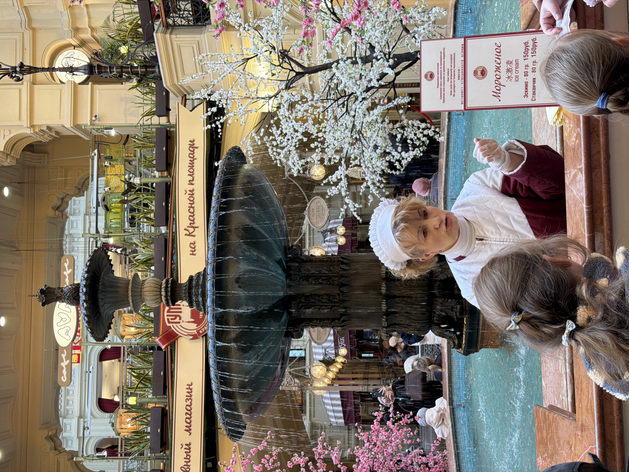 The image captures a scene inside the GUM (State Department Store) in Moscow, Russia. A grand, multi-tiered fountain takes center stage, adorned with floral decorations, including white and pink cherry blossoms. Adjacent to the fountain, a woman dressed in traditional attire, possibly a vendor, stands behind a counter. She is serving customers, presumably selling ice cream as indicated by the sign "Мороженое ice cream." The price list shows "Эскимо - 80 гр. 150руб." and "Стаканчик - 80 гр. 150руб." in Russian currency. There is a sign indicating that bank cards are temporarily not accepted. Patrons, including a young girl, are gathered around the counter, engaging in transactions. The interior of GUM, with its multiple levels and decorative lighting, provides a backdrop to the scene. The sign near the fountain reads "на Красной площади" which means "on Red Square."