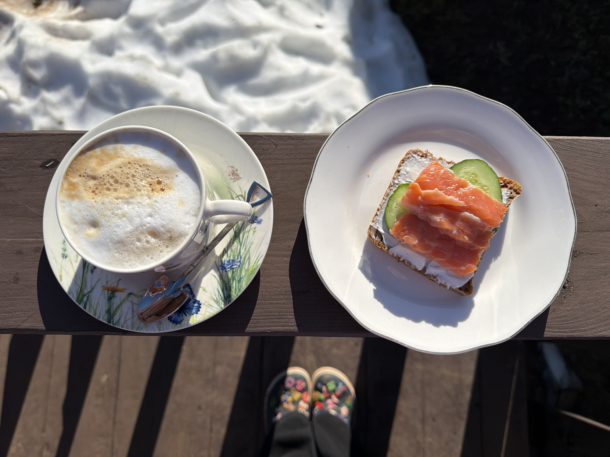 The image depicts a top-down view of a cappuccino and a salmon sandwich on a wooden surface, likely a balcony or deck. A portion of a snow-covered landscape is visible in the background, suggesting a cool climate.
On the left, a white cup with a floral pattern sits on a matching saucer, containing a cappuccino with frothy milk. A silver spoon rests on the saucer.
On the right, a white plate holds a sandwich made with dark bread, a white spread (likely cream cheese), salmon slices, and cucumber slices.
Below the plates, a pair of feet wearing dark pants and floral-patterned shoes are partially visible, indicating a person standing behind the camera.
The scene suggests an outdoor breakfast or snack on a wooden deck in a location with remaining snow. Based on the presence of the city of Chaikovsky in the prompt, it is inferred that the location is in Chaikovsky, Russia.