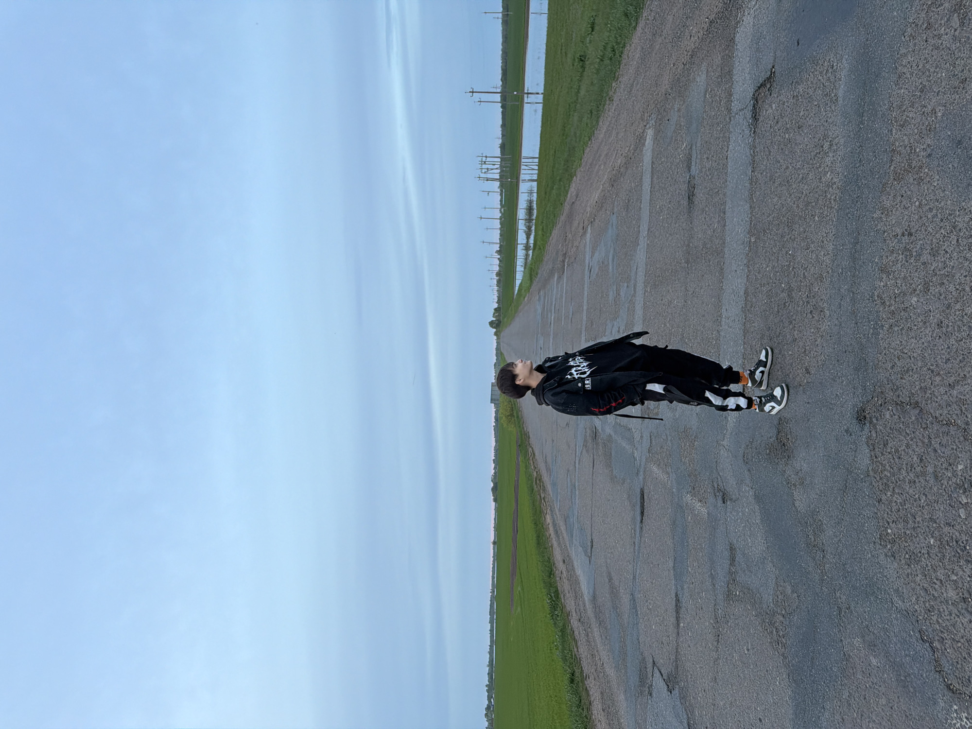 An individual is standing on a paved road that stretches into the distance, with fields and telephone poles on either side. The sky above is overcast. The road exhibits cracking and patching. The person is wearing dark clothing and distinctive black and white sneakers. They are looking upwards. The precise location within Azyakul', Russia is not discernible from the provided image.