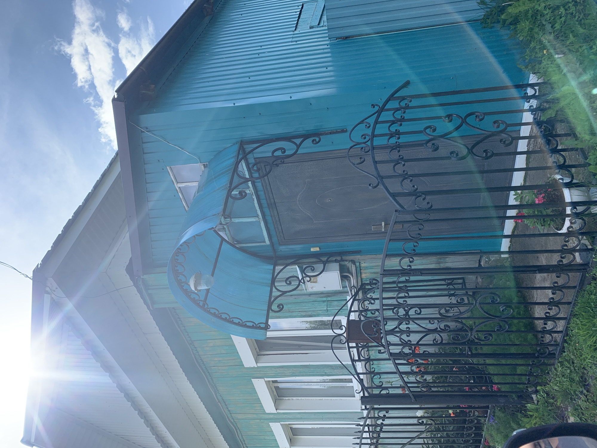 The image shows the exterior of a turquoise-colored wooden house with a dark metal door, situated behind a black wrought-iron fence. The house has several white-framed windows and a blue arched awning over the entrance. A light fixture is attached to the awning. A white electrical box is mounted on the wall next to the door. In front of the house, there is green grass and some potted plants with red and pink flowers. The sun is shining brightly, creating lens flares and highlighting the corrugated metal siding on the right side of the house. The sky above is blue with scattered white clouds. The house appears to be located in a rural or suburban area, possibly in the city of Barsai, Russia, given the context of the request.