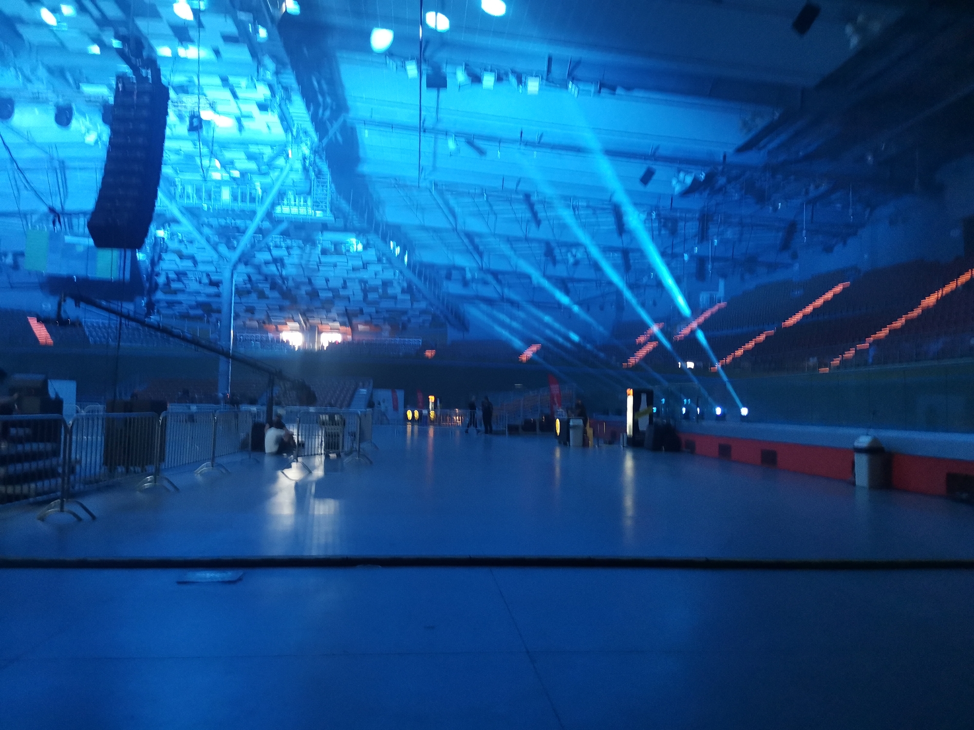 The image depicts the interior of a large, empty arena or concert hall in Plovdiv, Bulgaria, illuminated predominantly by blue light. The overhead ceiling structure is complex, featuring a network of trusses, beams, and numerous white circular light fixtures. Large, dark speaker arrays are suspended from the ceiling on the left side of the frame. Multiple strong blue spotlights project beams diagonally across the upper space towards the right. Tiers of unoccupied seating, dark in color, are visible along the right and rear perimeter of the venue, accented by horizontal lines of small orange lights, possibly marking steps or sections.
The main floor area, which appears to be polished and reflective, is a dark blue. In the mid-ground, several individuals, likely event staff or crew, are sparsely distributed across the floor, some standing, one seated near a section of silver metal crowd control barriers. These barriers create an enclosed area on the left side of the floor. Various indistinct pieces of equipment or stands are visible on the floor in the mid-ground. A horizontal black element runs across the lower portion of the foreground, potentially a cable run or floor transition. On the far right, a light-colored waste receptacle stands against a low red wall section. The overall impression is one of a venue either in preparation for an event or during tear-down, with a strong atmospheric blue lighting scheme.