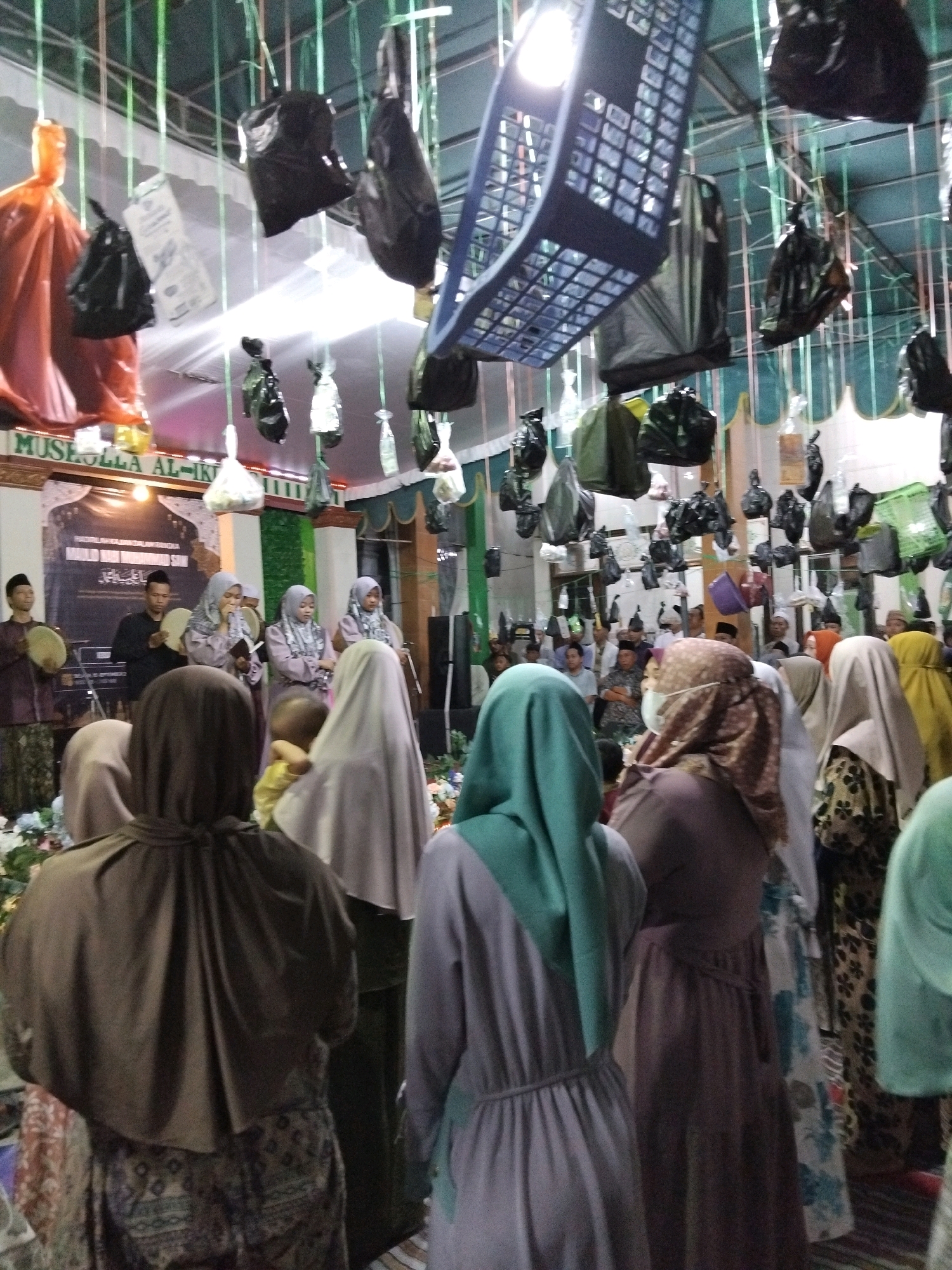 The image depicts an indoor gathering at the Musolla Al-Ikhlas in Kepuh Kiriman, Indonesia. In the foreground, several individuals are facing away from the camera, dressed in modest Islamic attire, including hijabs and long dresses. The central focus is a group of women in the middle ground, some of whom appear to be singing or reciting from books. To the left, two men are visible holding frame drums. The environment is decorated with hanging items, including black plastic bags, a blue basket-like structure, and what appear to be white bags. A banner with text is displayed on the back wall, partially obscured by the attendees. The overall atmosphere suggests a religious or cultural event taking place within a community space.
