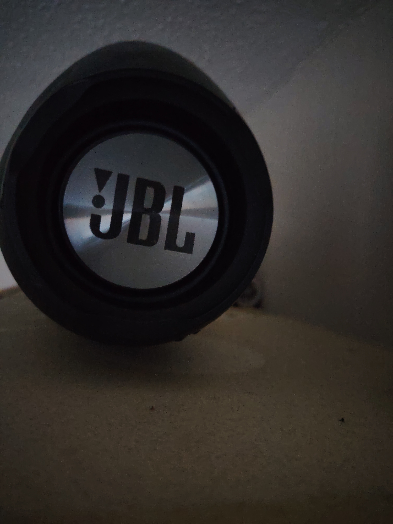 A close-up, low-angle shot shows the front of a black JBL speaker with a silver circular logo featuring the "JBL" brand name in stylized font. The speaker is angled upwards and to the left, resting on a textured, light brown surface, possibly a desk or shelf. The background is a dimly lit, textured gray surface, possibly a wall or ceiling. The image appears to be taken in a domestic setting in Huntingdonshire, United Kingdom. There are no people or animals visible in the scene.