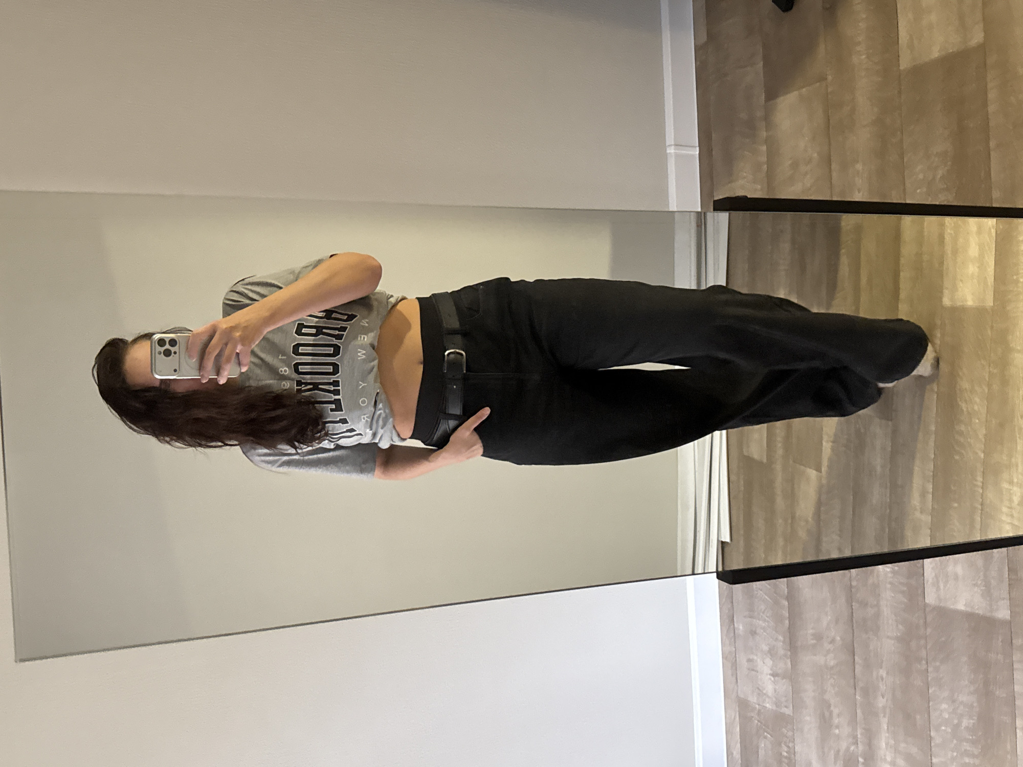 A young woman stands in front of a mirror, taking a photo with her phone. She is wearing a gray "Brooklyn" T-shirt, black pants, and a black belt. The background includes a plain wall and a wooden floor. The location is unknown.