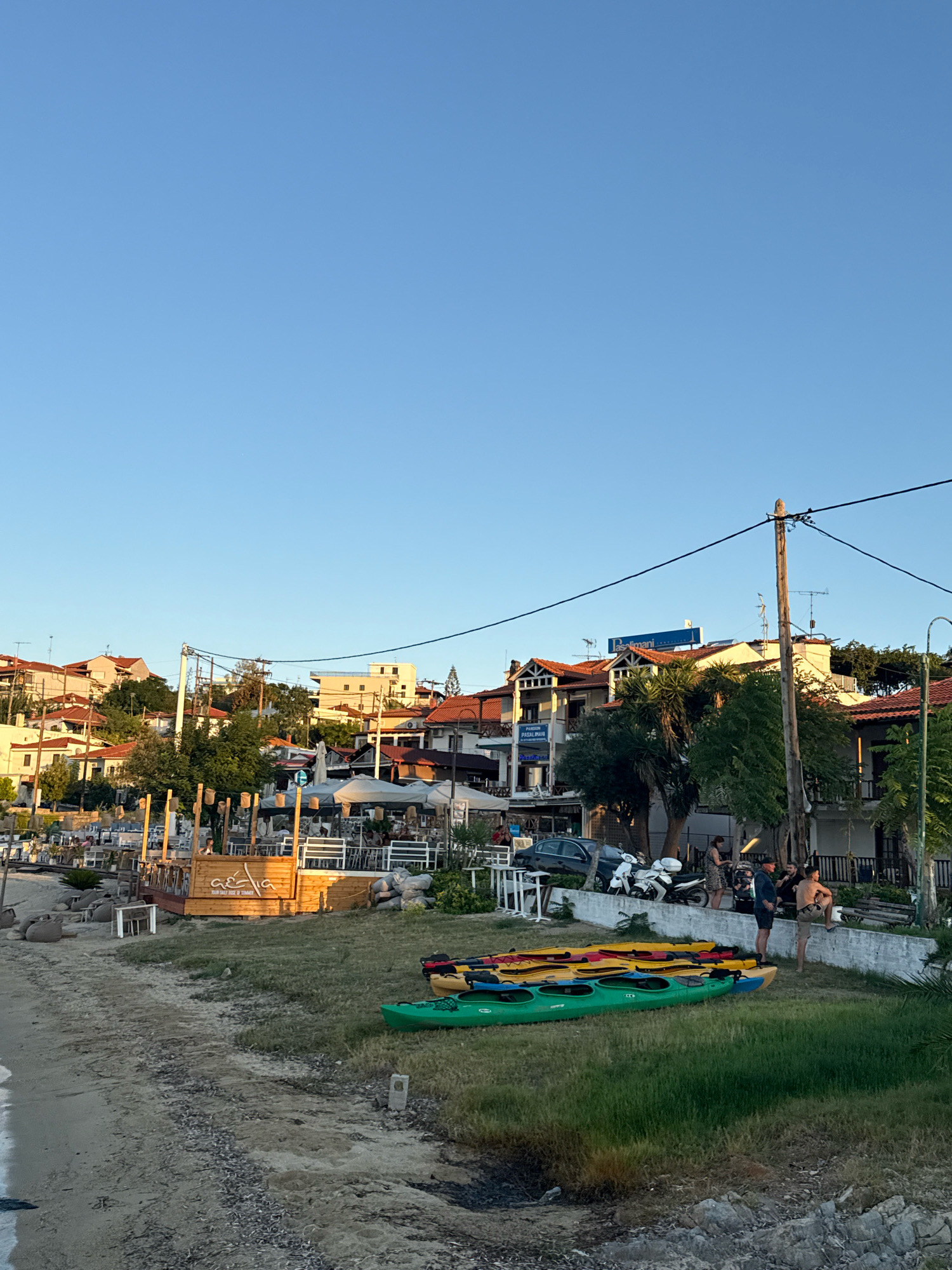 The image depicts a coastal scene in Ammouliani, Greece, with a clear blue sky overhead. In the foreground, a sandy beach transitions to a grassy area where several kayaks are stacked. These kayaks are in various colors, including green, yellow, and red. To the right of the kayaks, a group of four individuals are standing and interacting; one person is seated, appearing to be stretching.
Behind the kayaks and grassy area, a section of the town is visible. Buildings with red-tiled roofs are clustered on a gentle slope. Along the waterfront, there are outdoor seating areas for what appear to be cafes or restaurants, some with umbrellas for shade. Wooden structures and furniture are present in these dining areas. Further back, more buildings are visible, as well as trees and utility poles with electrical wires. A dark-colored car and two scooters are parked near the establishments. The overall impression is a seaside town during the daytime, likely in the late afternoon or early evening given the lighting.