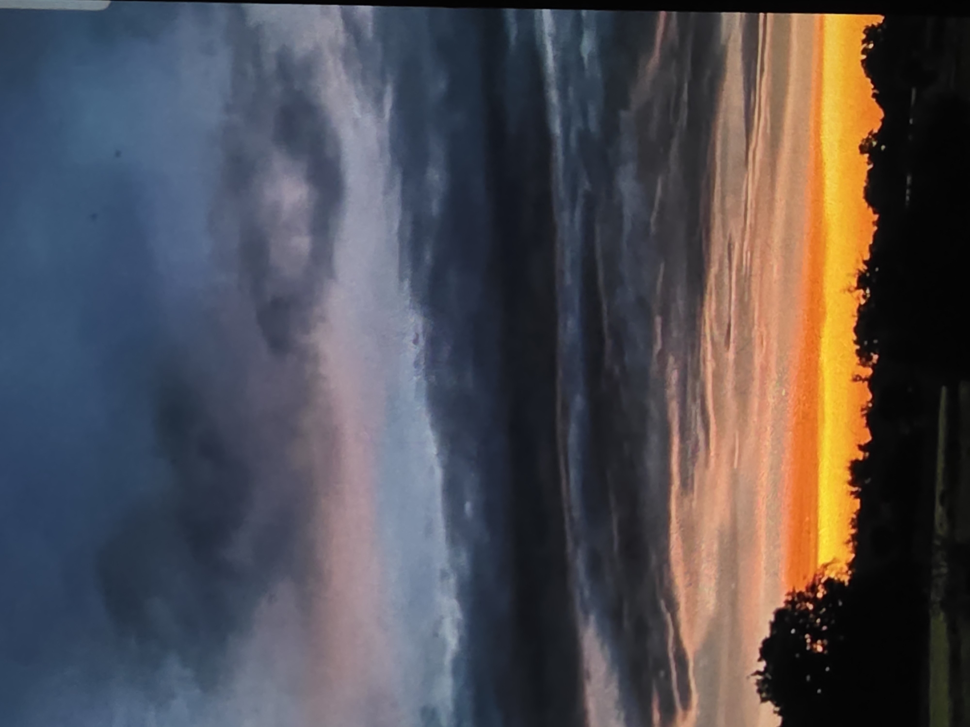 This is a vertical, wide-angle photograph of a dramatic sunset over a rural landscape. The sky is filled with thick, dark clouds in various shades of gray and blue, with a bright band of orange and yellow light emanating from the horizon. The clouds are layered, with some appearing to be illuminated from behind by the setting sun. In the foreground, the silhouettes of trees and a low horizon line are visible. The setting is a rural area, likely on the outskirts of Dubno, Ukraine. There are no people or animals visible in the image. The overall mood is one of dramatic natural beauty.