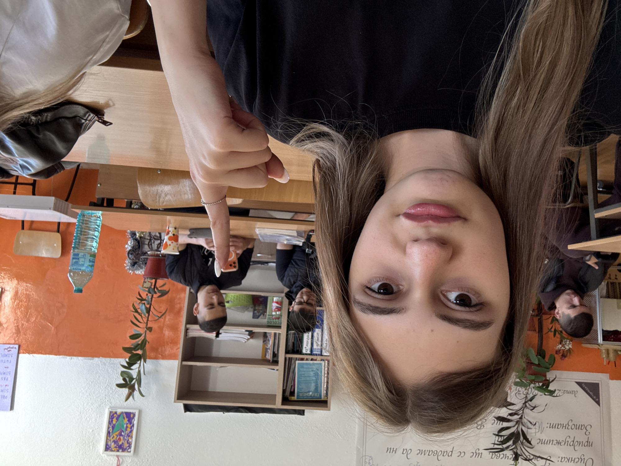 A selfie taken indoors, likely in a classroom, features a young woman with light brown hair. She wears a black top and a ring on her index finger. Behind her, seated at desks, are two boys, one wearing a black shirt and using a phone, and the other wearing a black jacket. A bookshelf with books and a plant are visible. The wall is painted orange and white, with posters and text written in Cyrillic. Based on the Cyrillic text and available information, the location may be Panagyurishte, Bulgaria.