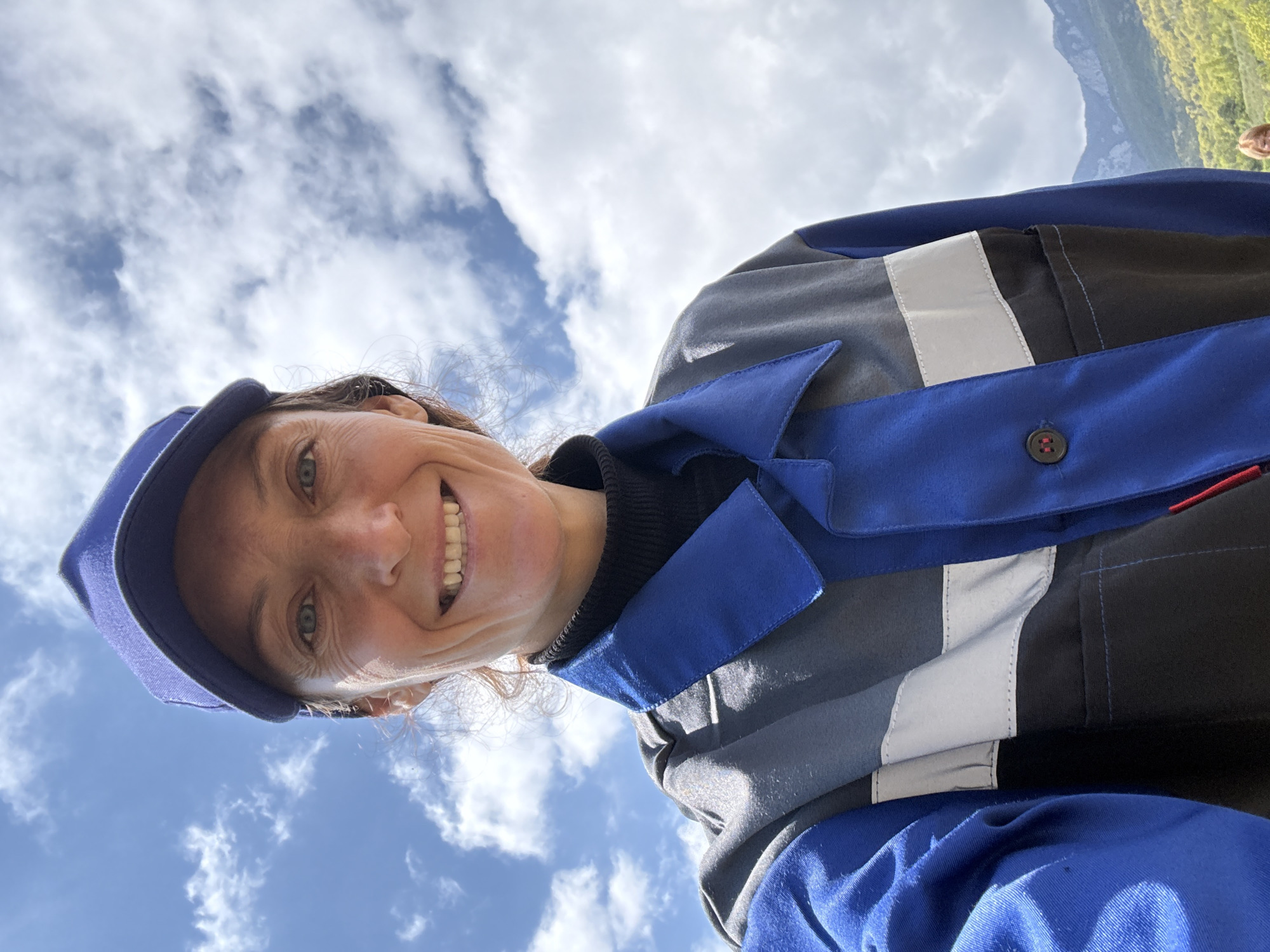 A low-angle, slightly rotated selfie shows a smiling woman wearing a blue cap and a multi-colored jacket. The jacket features blue, grey, white, and black panels. The background consists of a blue sky with scattered white clouds. In the upper right corner, a small section of green foliage suggests a natural environment. The location is not discernible as Pushkino, Ukraine from the visual information provided.