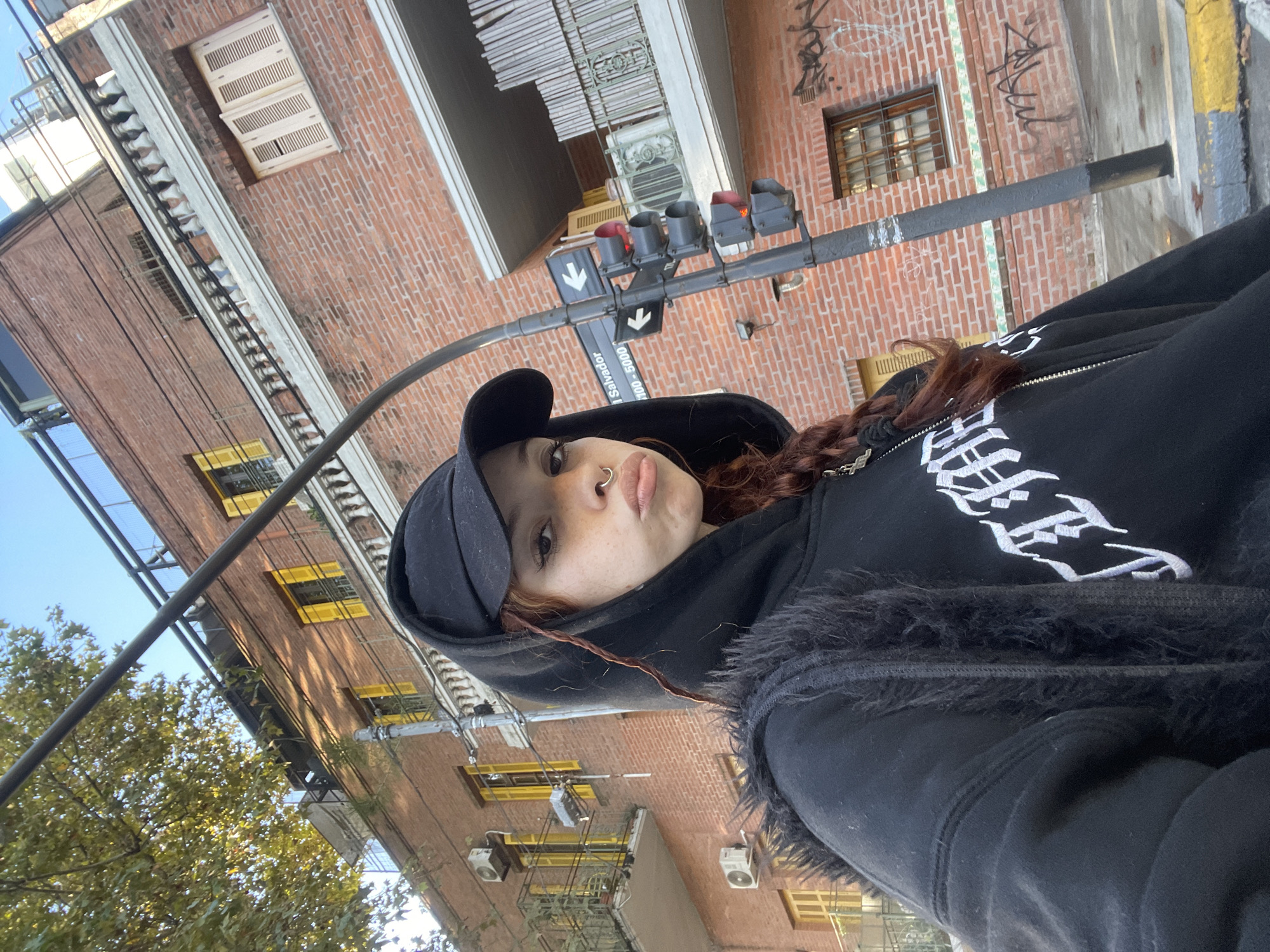 This image, captured at a low angle and rotated 90 degrees clockwise, depicts a young person posing for a photograph in an urban environment. The main subject is a person with reddish-brown hair styled in braids, wearing a black baseball cap and a black zippered hoodie. The hoodie features stylized white text embroidery on the front and a furry black trim along the zipper. A nose ring is visible on the subject's face, which holds a neutral expression with subtly pursed lips.
Behind the person, the scene is dominated by multi-story brick buildings under a clear daytime sky. The buildings feature numerous windows, some framed in bright yellow, and external air conditioning units. A black street pole extends across the frame, supporting a traffic light displaying red signals and a street sign. The sign, rotated with the image, clearly displays "Salvador" and the number range "100 - 5000," along with downward-pointing arrows, strongly indicating a street in Buenos Aires, Argentina. Illegible graffiti is visible on some of the brick walls. To the left, green trees are abundant, suggesting proximity to a park or tree-lined street. Wires and cables crisscross the upper portion of the scene. A yellow and grey painted curb is partially visible at the bottom.