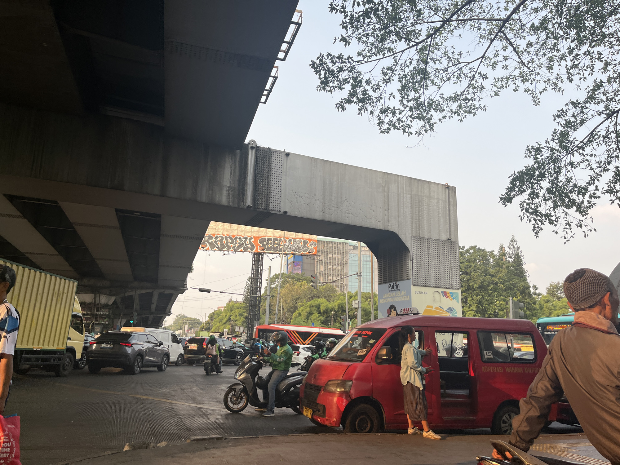 This image captures a bustling urban scene in East Jakarta, Indonesia, likely during daylight hours given the illumination and sky conditions. The foreground features a red minivan with its rear passenger door open, and a woman in a light blue shirt and striped skirt is standing next to it, facing the open door. To the right of the minivan, a person wearing a dark jacket and a knitted cap is partially visible, facing away from the camera and towards the traffic.
The midground reveals a busy street with various vehicles. A motorcycle rider in a green jacket is on a scooter, looking at their phone. Behind them, several other motorcycles and cars are present, indicating traffic congestion. Further back, a white van and a dark grey SUV are seen, along with a large yellow truck to the far left. In the background, a long red bus is visible, as are trees and tall buildings.
The scene is dominated by overhead infrastructure, likely an elevated road or bridge, casting shadows on the street below. Graffiti is visible on a large banner or wall under the elevated structure. The overall environment suggests a typical city street with significant traffic flow and urban elements. The sky is mostly clear, with some branches of a leafy tree extending into the upper right portion of the frame.