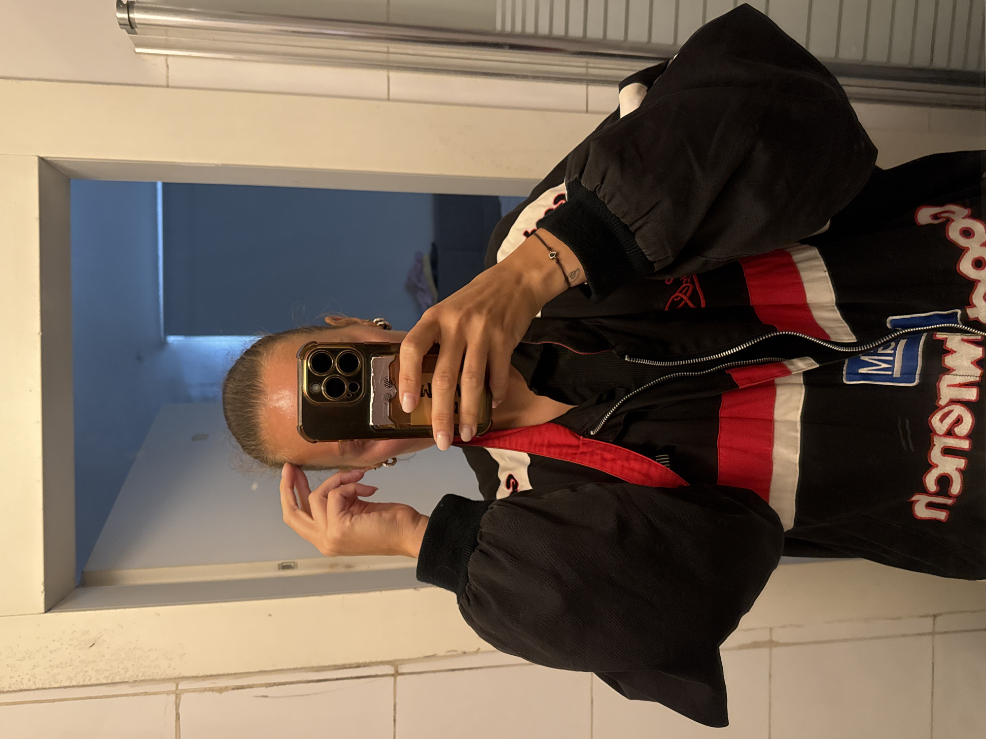 A person is taking a selfie in a bathroom, likely in Tel-Aviv, Israel. The person is holding a gold-colored smartphone. The background features a white wall and a mirror reflecting an interior space with a door. The person is wearing a black racing-style jacket with red and white stripes and lettering. The bathroom has white tiles. A bracelet is visible on their wrist. The overall scene is indoors.