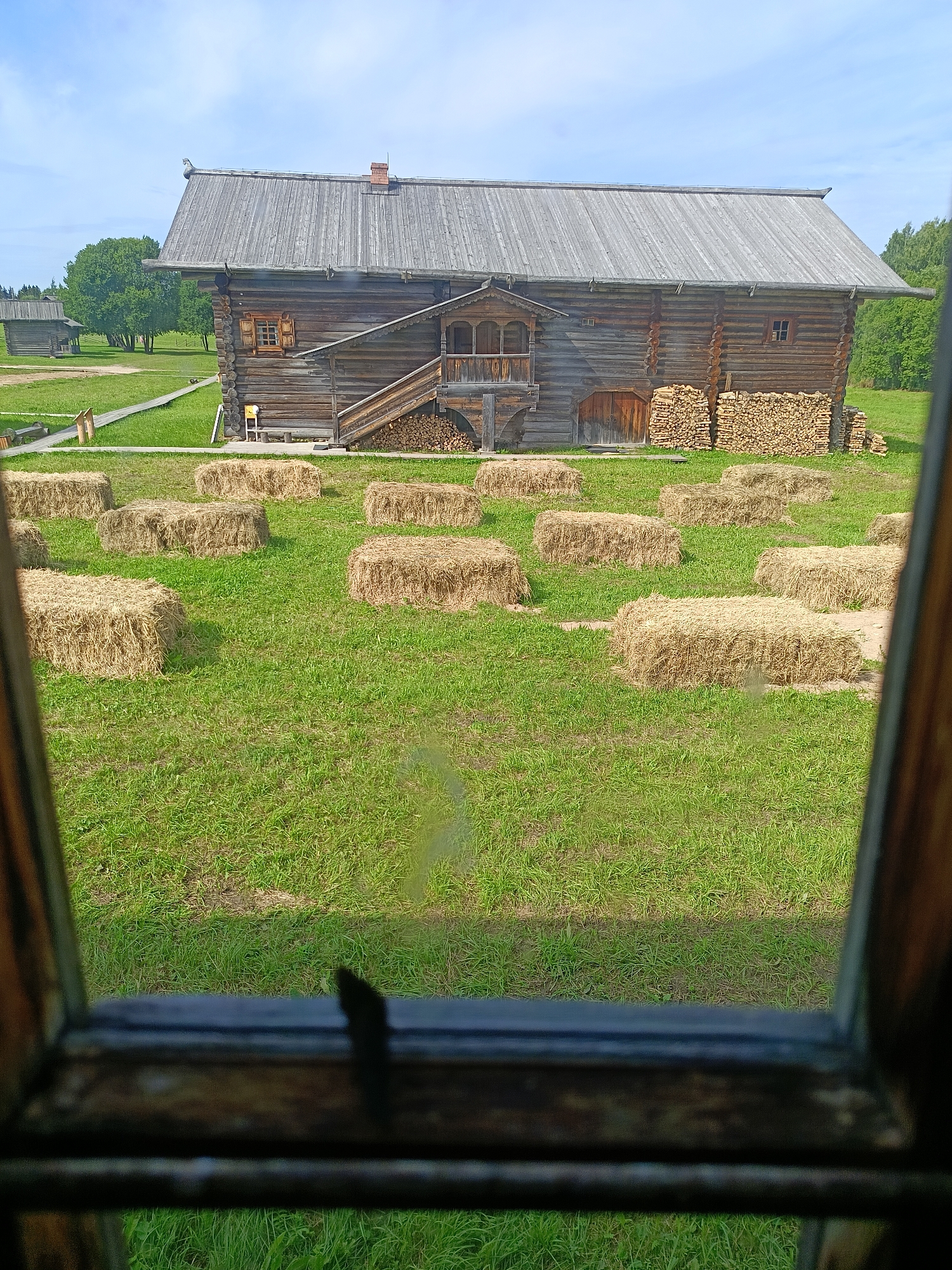 An outdoor, eye-level, medium shot captures a rustic wooden barn in the background, with a green grassy field in the foreground. The barn, constructed from large logs, features a steeply pitched wooden roof, a covered porch with a staircase leading up to it, and several small windows. Stacks of firewood are piled against the side of the barn. In front of the barn, the field is dotted with numerous rectangular bales of hay, arranged in a scattered pattern. The foreground of the image is dominated by a blurred wooden window frame, suggesting the photo was taken from inside a building looking out. The sky above is a pale blue with scattered white clouds. The location is described as Unknown, Unknown.