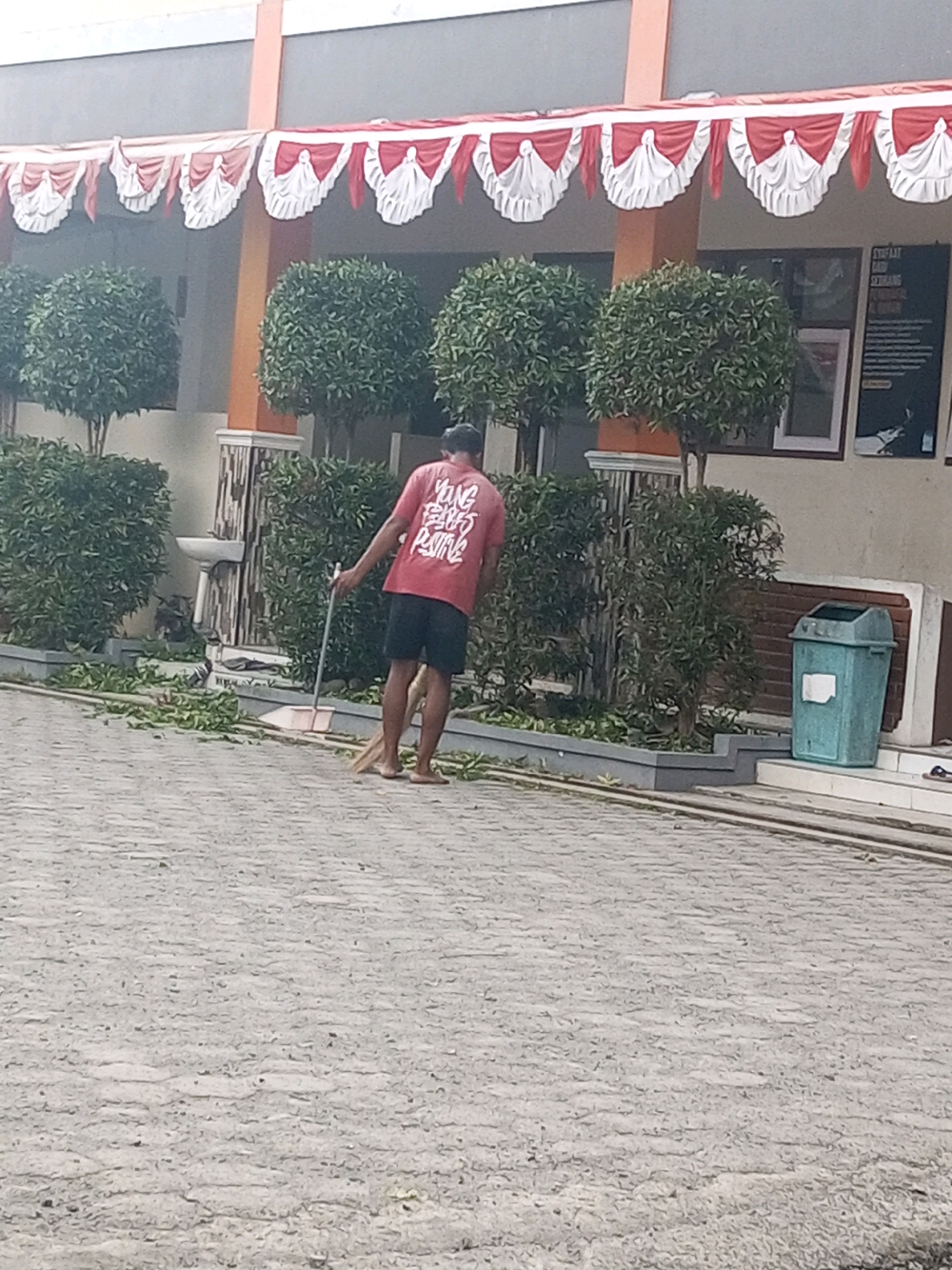 A man in a red t-shirt and black shorts is sweeping leaves on a paved area in front of a building. The building has decorative red and white fabric draped along its facade. Several potted trees are visible in front of the building, and a teal trash can is to the right of the frame. The scene appears to be outdoors, likely in a public space like a school or office building.