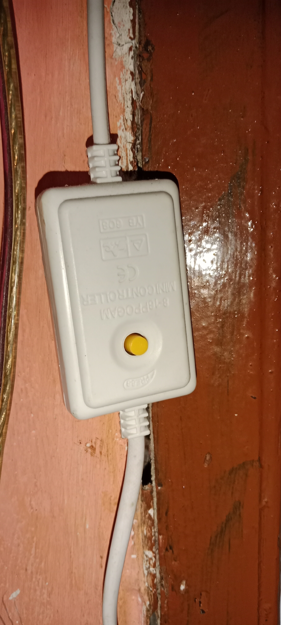 A white plastic mini-controller box is mounted on a coral-colored wall. The box has a small yellow button in the center. There are several markings on the box, including "YB 609", a symbol of a person with an "X" over them, an equilateral triangle with an exclamation mark, and "CE". Text below the button reads "B-18PPOGAM MINI CONTROLLER". A white cable enters the top of the box, and another white cable exits the bottom. To the left of the box are several wires with a braided metallic exterior. The wall is weathered, with peeling paint and a darker, textured surface on the right side. The location is identified as Subang, Indonesia.