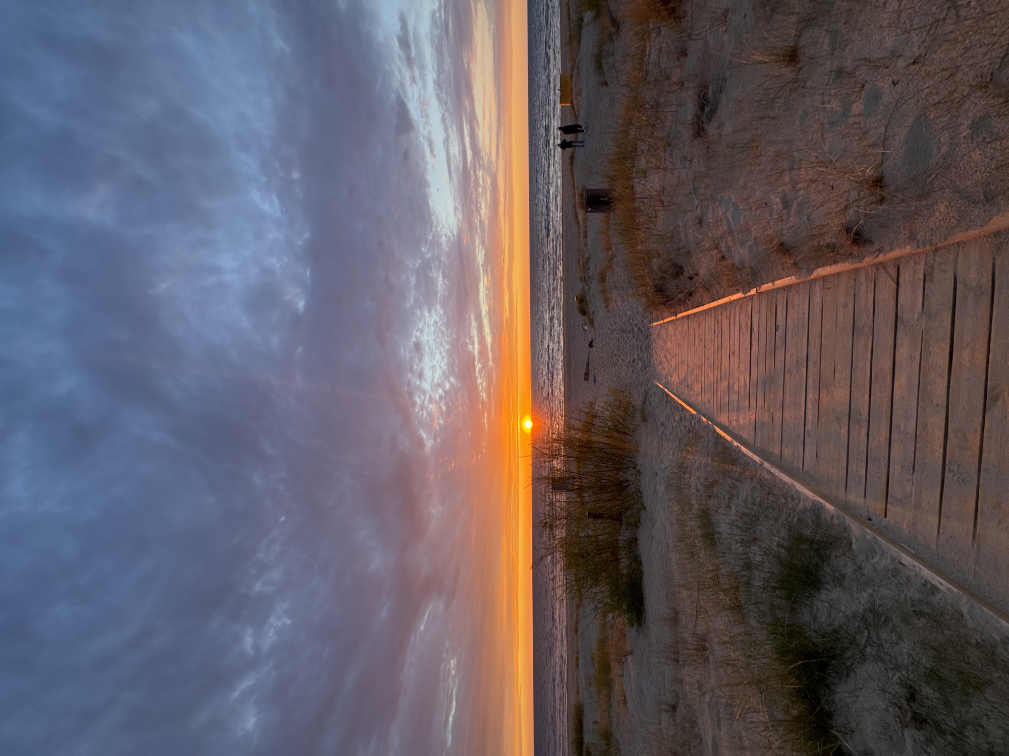 The image depicts a sunset at a beach in Divezeri, Latvia. The sun is setting over the water, with the sky displaying shades of orange, pink, and blue. There are clouds in the sky. A wooden walkway extends towards the beach from the right side of the image. Sand and dune vegetation line the walkway. Two people can be seen standing near the shore in the distance. The water is calm.