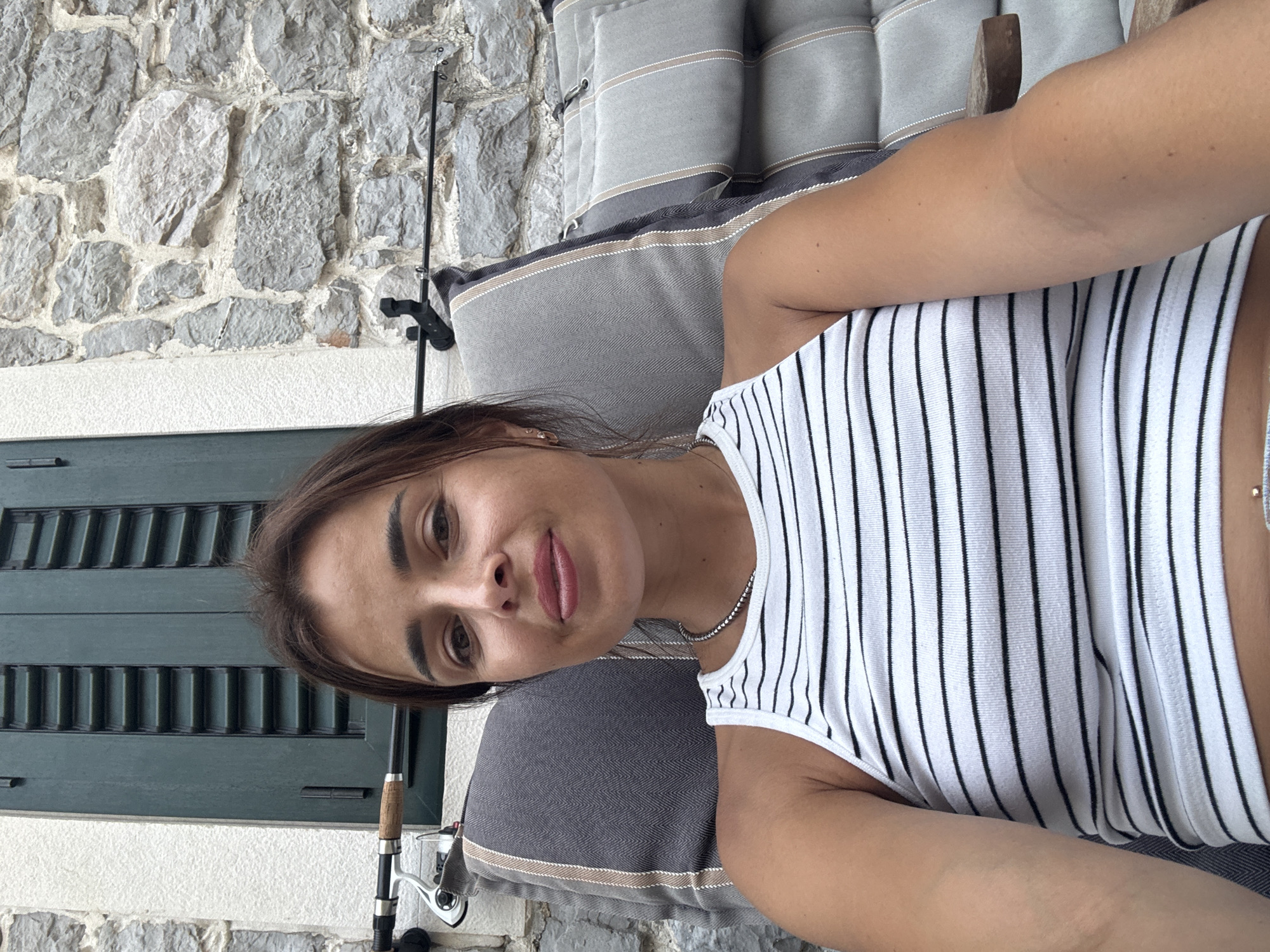 A medium shot captures a woman with brown hair and light skin, looking at the camera while sitting on a gray outdoor chair with striped cushions. She is wearing a white tank top with thin black horizontal stripes and a silver necklace. Behind her, a stone wall is visible, with a green shuttered window to her left and two fishing rods mounted on the wall behind her.