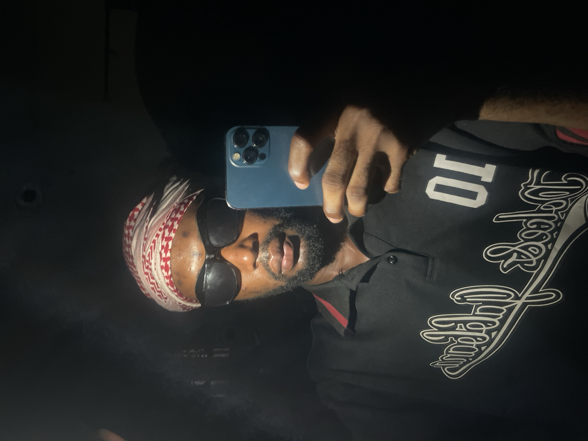 A dark-skinned man is holding a blue smartphone up to his face, presumably taking a selfie. He is wearing a black collared shirt with a white design and the number "10" on it, a red and white patterned head covering, and dark sunglasses. The background is dark and difficult to discern, though there appears to be a dim light source. The location is presumed to be Lokoja, Nigeria. There is no further information to infer about the environment.