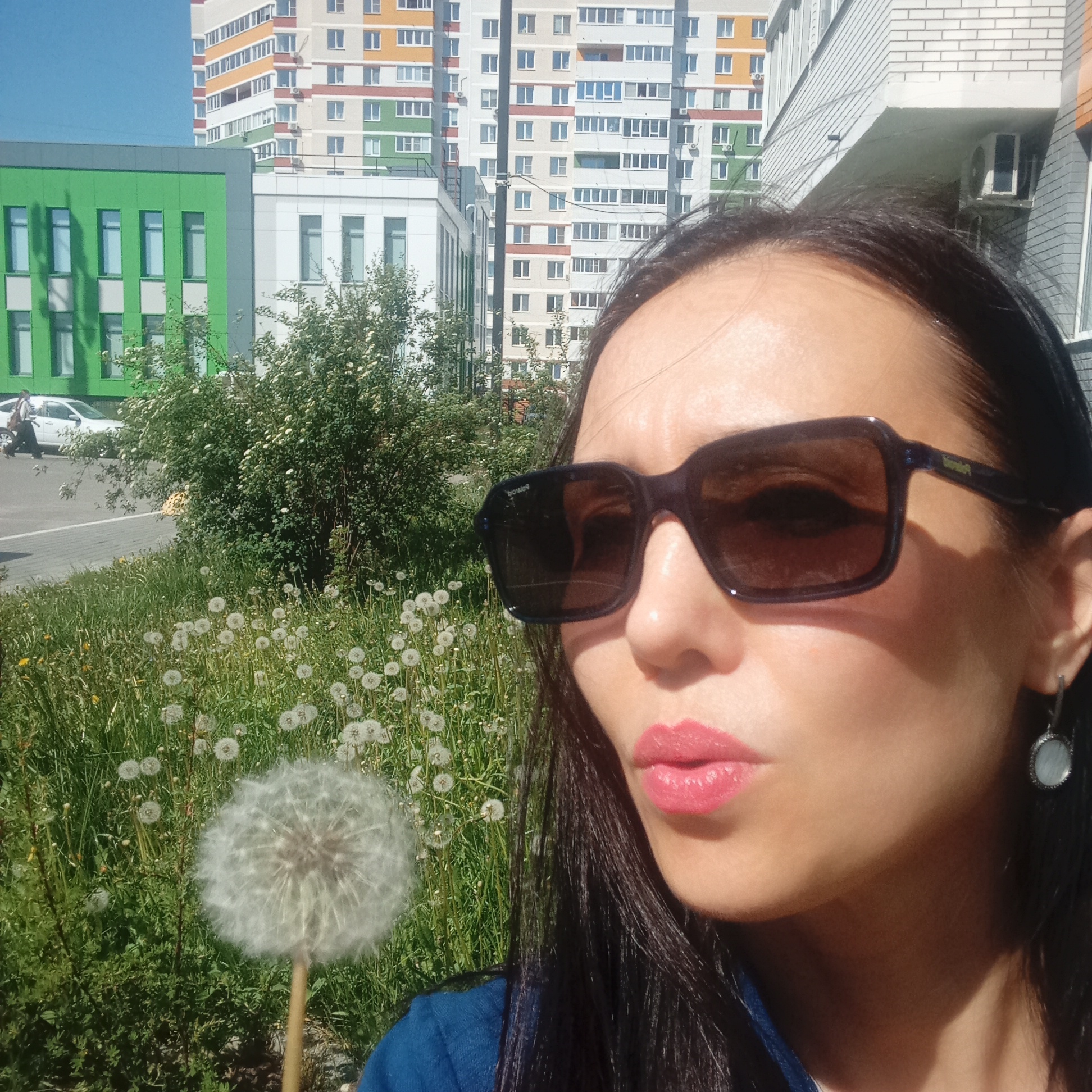 A woman with long, dark hair is holding a dandelion and preparing to blow on it. She is wearing dark sunglasses and pink lipstick. In the background are several multi-story buildings, including one with a green and gray facade, and another with orange, white, and green horizontal stripes. The buildings are typical of urban architecture. There is a patch of grass and dandelions in the foreground as well. The location appears to be in an urban or suburban area. The location is Izhevsk, Russia.
