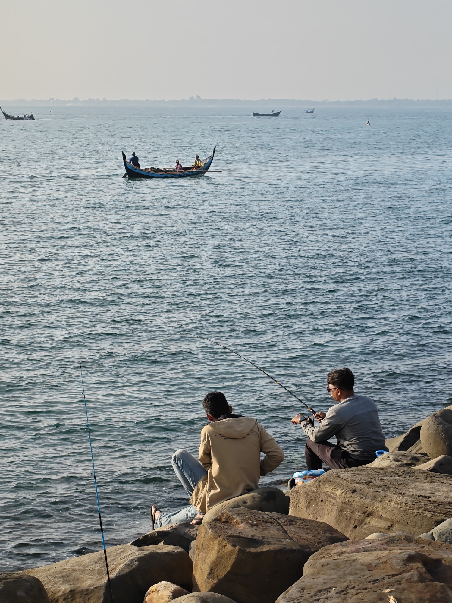 The image depicts a serene coastal scene in Lhokseumawe, Indonesia, likely during the late afternoon or early morning hours, indicated by the soft, warm light. In the foreground, two men are seated on large, textured rocks at the water's edge, engaged in fishing. The man on the left wears a light brown hooded jacket and light blue jeans, with a distinctive blue fishing rod propped upright beside him. The man on the right, dressed in a gray long-sleeved shirt and dark pants, actively holds his fishing rod, which has a black and gold reel, looking out towards the sea. A small blue and white cooler or bag rests on the rocks next to him.
The calm, rippling blue-green water dominates the mid-ground, where a traditional wooden fishing boat, painted black and blue with distinctive pointed ends, is seen with at least four people onboard. One individual appears to be at the stern, potentially steering or rowing. In the distance, several other boats are scattered across the vast expanse of water, appearing as dark silhouettes against the bright, slightly hazy sky. The horizon is a faint, light gray line where the sea meets the sky, suggesting a clear yet somewhat misty atmosphere. No visible text is present in the image. The overall scene conveys a sense of peaceful leisure and traditional maritime activity.