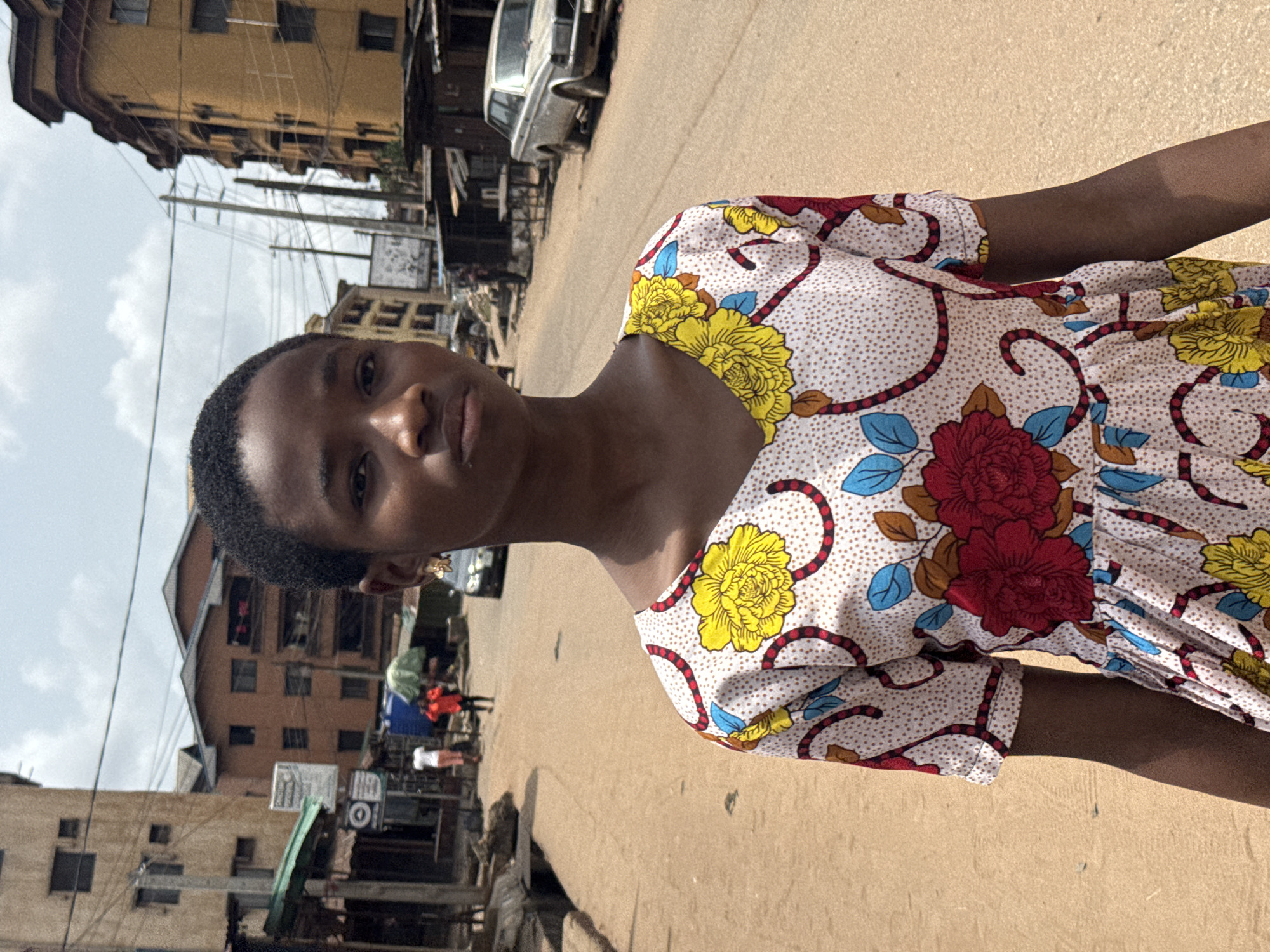 A young woman with short, dark hair stands facing the camera in what appears to be Onitsha, Nigeria. She is wearing a floral dress with red, yellow, and blue flowers on a white background. The street is unpaved and dusty. Buildings of various heights are visible on both sides of the street, with electrical wires crisscrossing the sky. A car is parked in the background. There are signs of commerce with stalls and people visible in the distance.