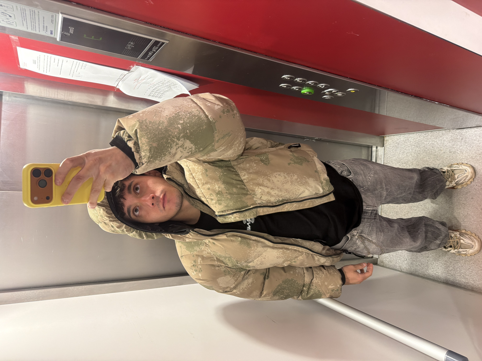 The image displays a young man taking a selfie inside an elevator, likely reflecting off a polished metallic surface. He is seen from the chest up, holding a bright yellow smartphone in his right hand to capture the photo. He wears a camouflage-patterned puffer jacket with a hood, a black t-shirt, faded grey jeans, and light-colored chunky sneakers. His expression is neutral as he looks directly at the camera.
The elevator interior features distinctive red and white striped panels on the upper section of the walls. Below these, a metallic panel houses a series of round elevator buttons, one of which is illuminated green, indicating a selected floor. On this control panel, text is visible, indicating a weight limit of "2020 kg" and a passenger capacity of "14/24 personas." A digital display shows what appears to be "000N". Several crumpled papers with largely illegible text are taped to the wall adjacent to the control panel, possibly notices or instructions. A horizontal metal handrail runs along the lower wall.
The setting is an indoor space, specifically an elevator, in what appears to be a modern building, situated in Sofia, Bulgaria. The image was taken from a slightly low angle. A partial word, "VE," is visible on the man's black t-shirt.