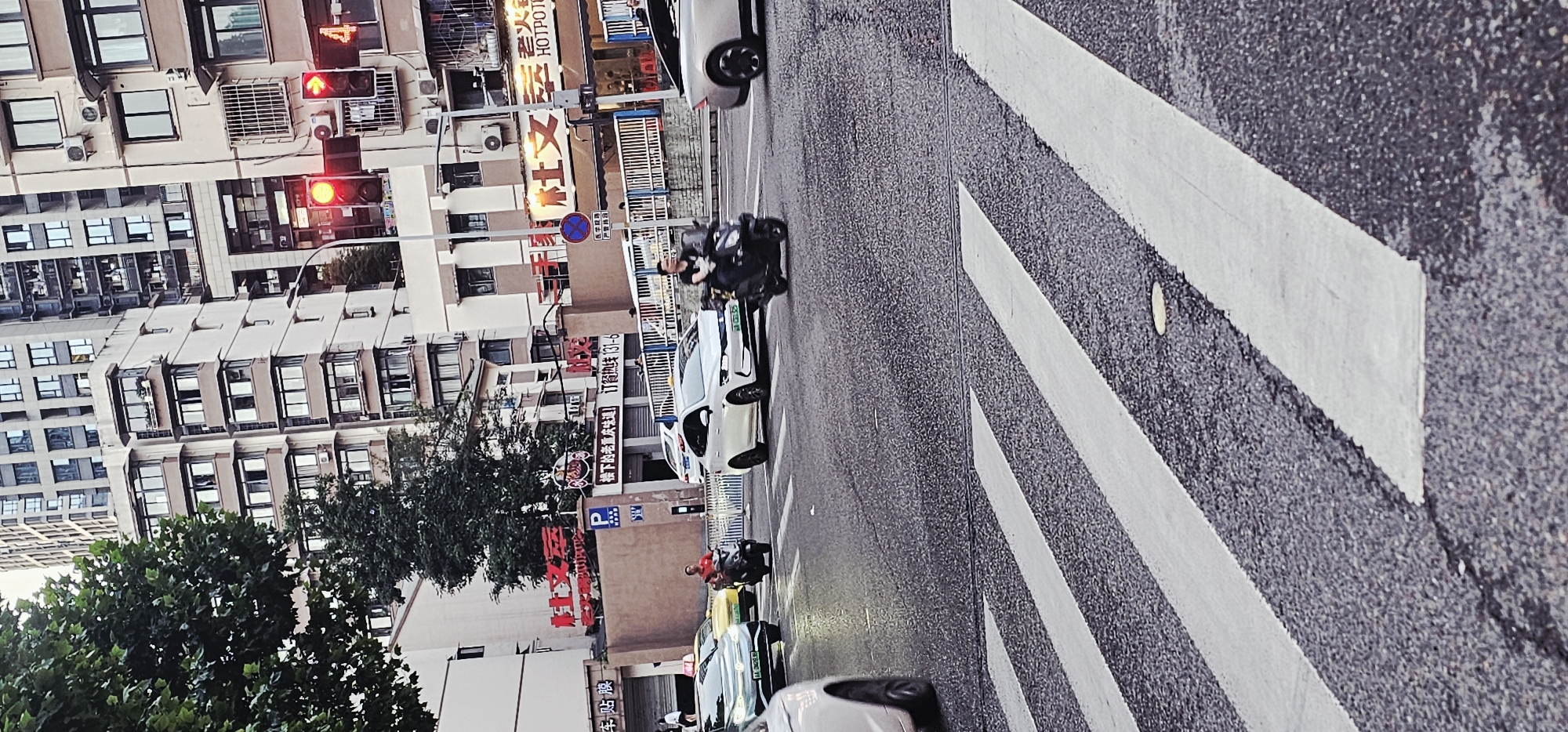 The image shows a street scene in Yuzhong District, China, featuring buildings, street signs, vehicles, and people. A crosswalk stretches across the asphalt roadway. Traffic lights are visible, showing red indications. Numerous vehicles are parked along the side of the road, including cars and motorbikes. Signs written in Chinese characters advertise restaurants and services. Pedestrians and people on motorbikes are present on the street. A large tree grows beside one of the buildings. The overall impression is of a bustling urban setting.