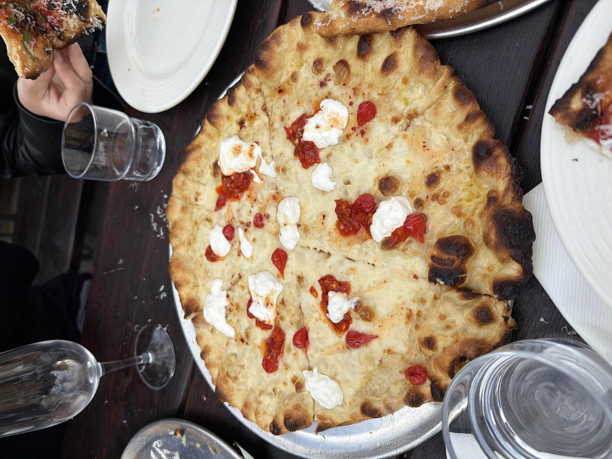 The image captures a tabletop scene, seemingly in a restaurant setting. A pizza with a slightly charred crust is the central element, topped with white dollops of what appears to be cheese and red tomato pieces. The pizza sits on a round, metal tray. Around the pizza, there are signs of dining: a partial view of a white plate with a slice of pizza, another white plate in the background, a glass lying on its side, another tall stemmed glass, and a short cylindrical glass. A hand holding a slice of pizza is visible in the upper left. The tabletop appears to be made of dark wood. The scene is well-lit, suggesting daytime or bright indoor lighting. While the exact location in London cannot be determined from the image, the scene suggests a contemporary dining environment.