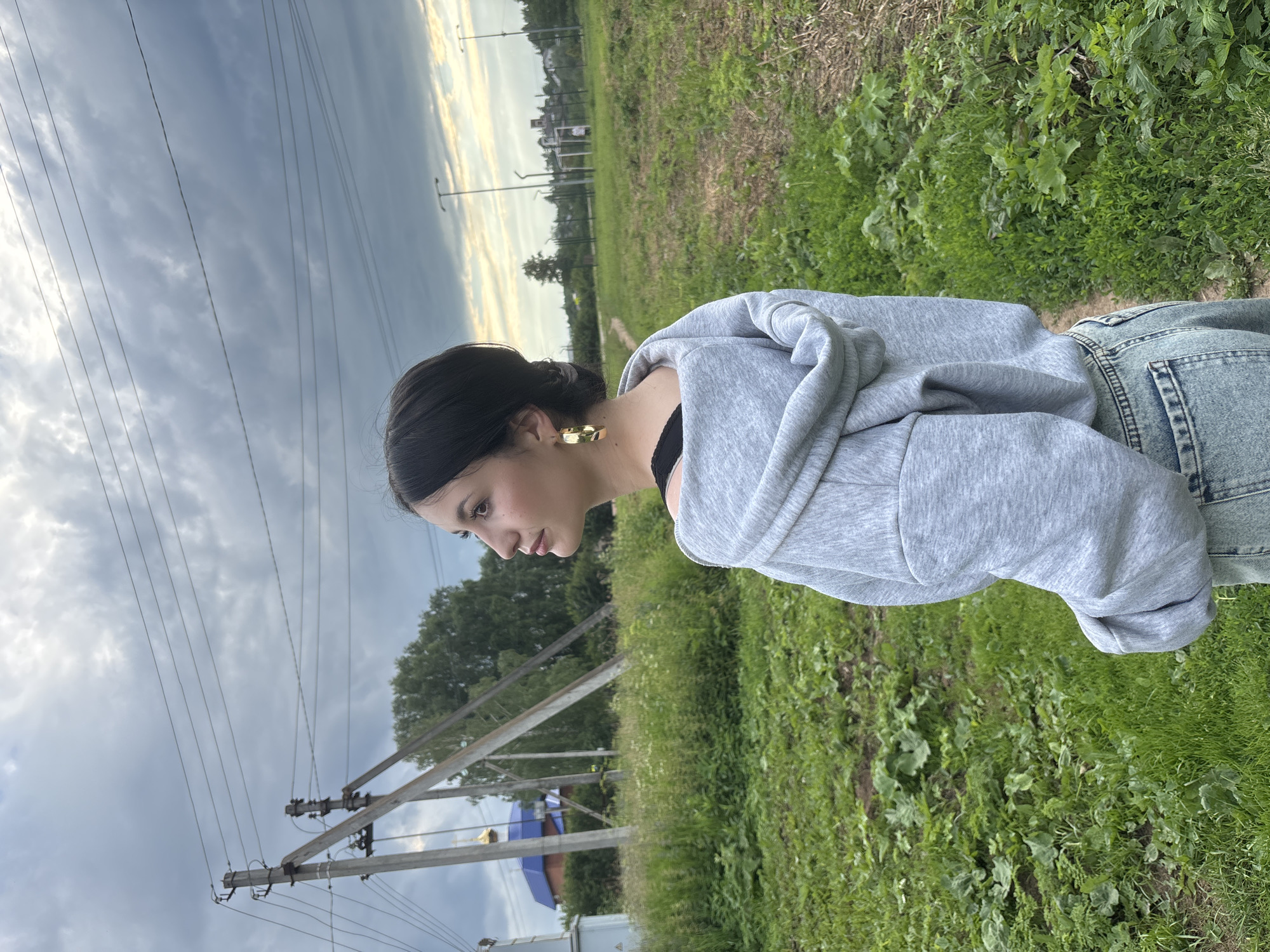 A young woman with dark hair and large gold hoop earrings is standing in a grassy field, looking to her left. She is wearing a grey hooded sweatshirt and light-wash denim jeans. In the background, there are power lines, a church with a blue dome, trees, and a chain-link fence with some structures beyond it. The sky is cloudy and grey, suggesting overcast weather. The location appears to be the city of Fokino, Russia.