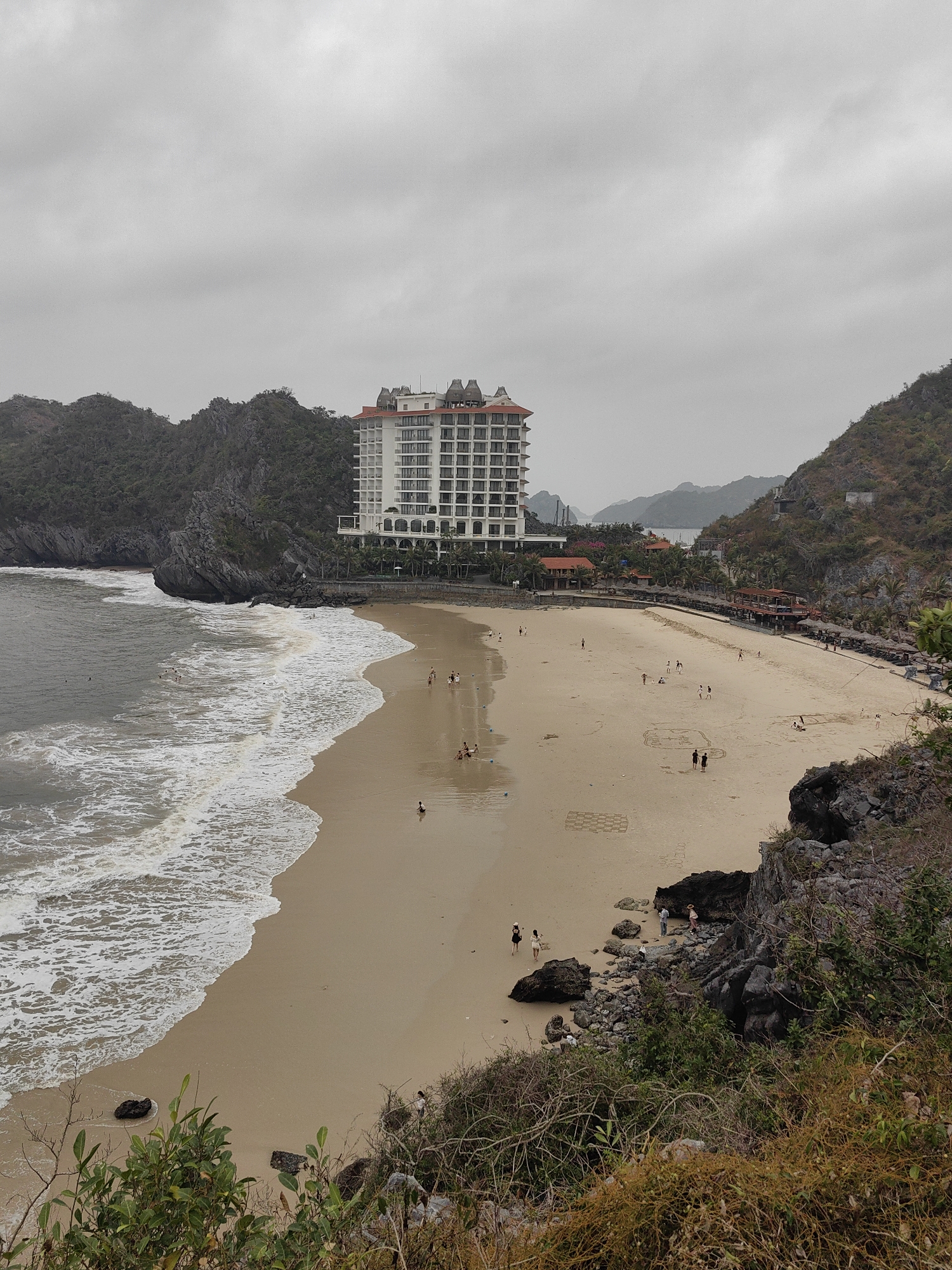 Captured from an elevated vantage point, the image showcases a beach scene in the city of Hải Phòng, Vietnam. A wide sandy beach stretches across the frame, lined by the ocean on one side and buildings and rocky terrain on the other. The ocean is rough with foamy waves breaking onto the shore. Several people are scattered across the beach; some are in the water, some are walking along the shore, and others are gathered in groups. A large, white multi-story hotel is situated at the edge of the beach, abutting rocky cliffs. Adjacent to the hotel are smaller structures with red roofs. The sky overhead is overcast, contributing to the subdued lighting of the scene. The foreground features vegetation, including plants and dry grass, growing on the rocky hillside where the photograph was taken.