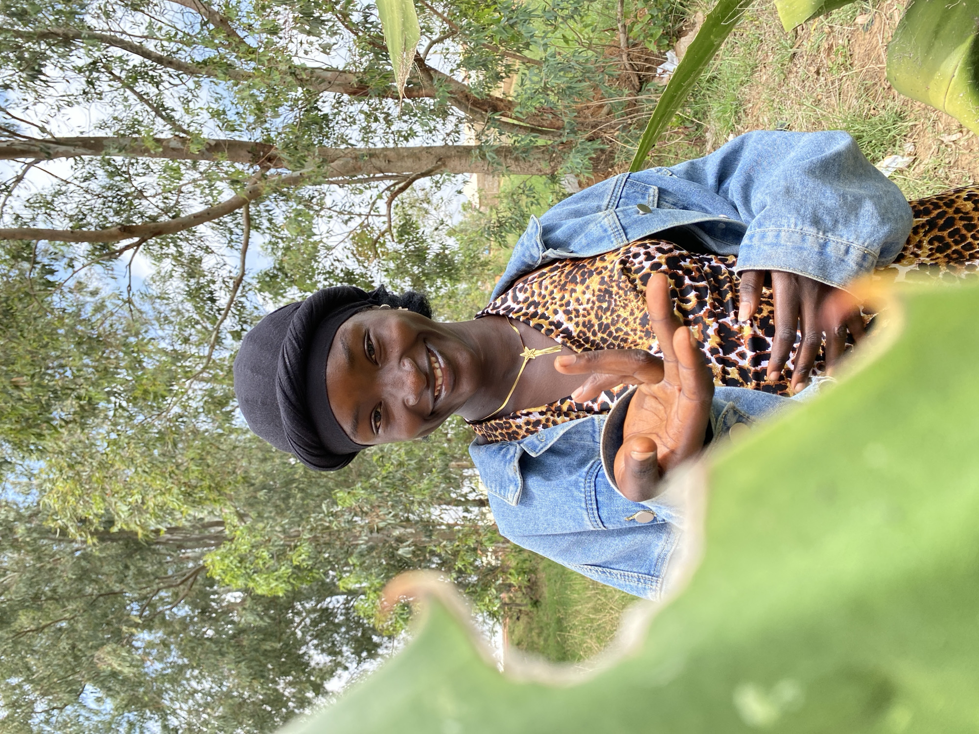 A smiling person wearing a black head covering, a leopard-print top, and a denim jacket is positioned outdoors. The person is making a peace sign with their right hand. The environment features trees and greenery, some of which are blurred in the foreground. The location is unknown; it cannot be determined to be in Durimi, Nigeria.