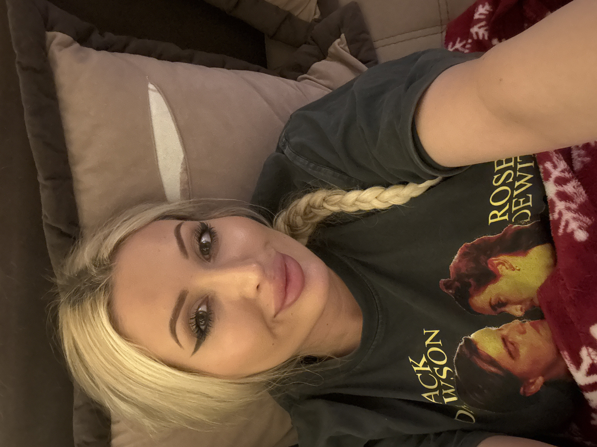An adult female with blonde hair, styled in a braid over her right shoulder, is lying down and looking at the camera. She is wearing a dark t-shirt with images of Jack Dawson and Rose DeWitt from the movie Titanic printed on it, along with the text "JACK DAWSON" and "ROSE DEWITT". She is also covered by a red blanket with white snowflake patterns. The background consists of pillows. The image is a selfie taken from a close-up perspective.